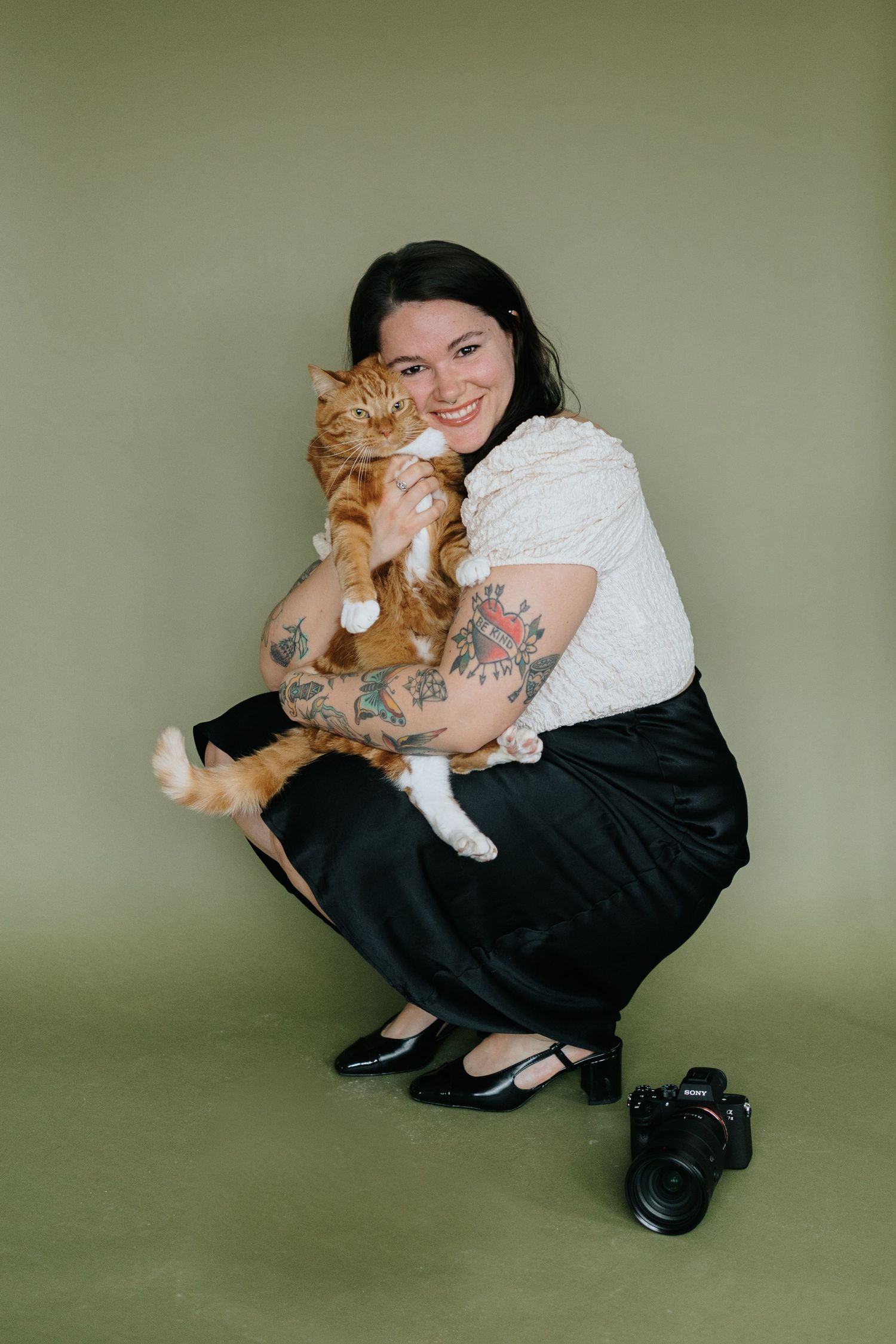 A ginger cat cuddling with someone in casual attire against a sage green background.