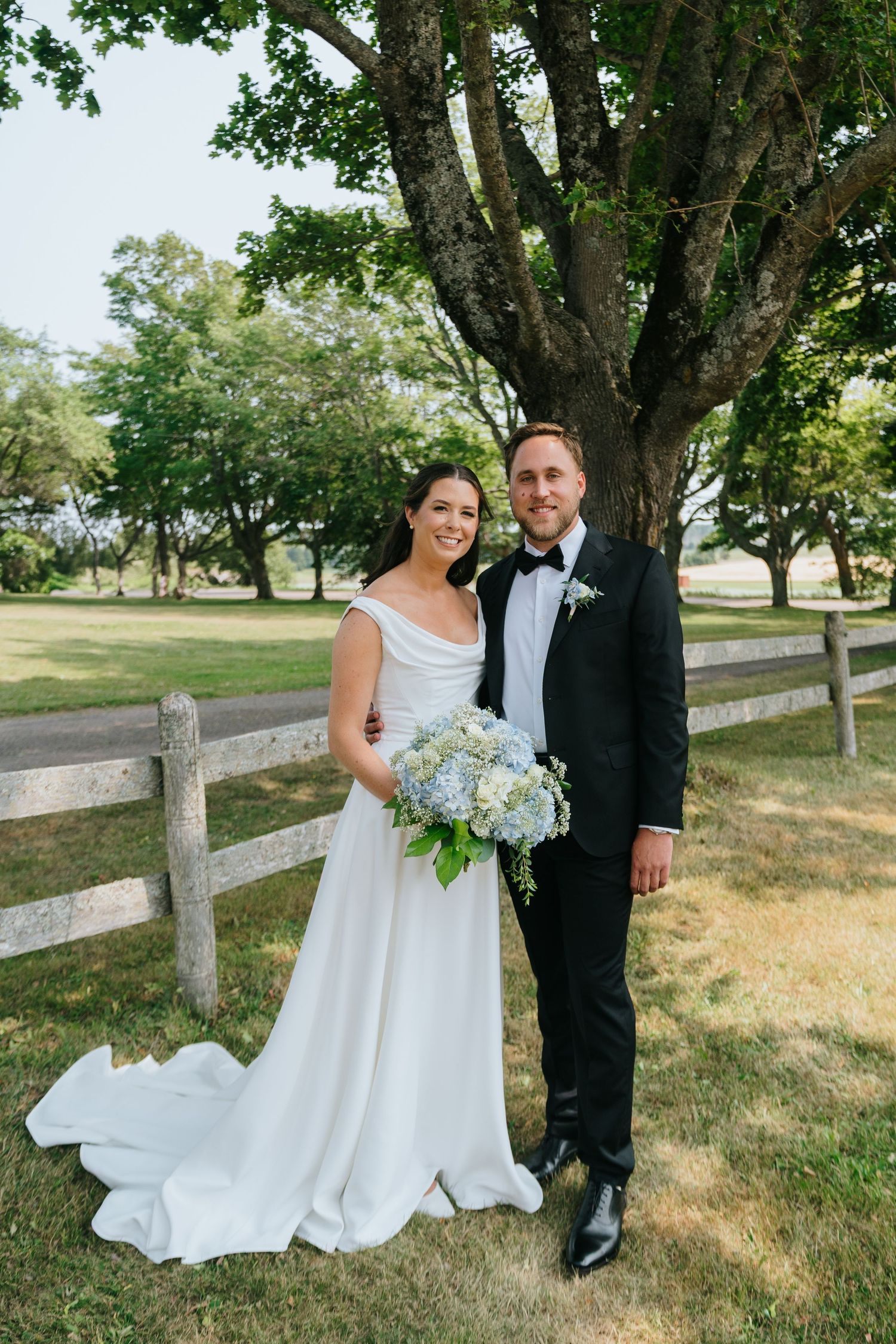 A couple poses for a formal portrait in front of a rustic fence and large tree.