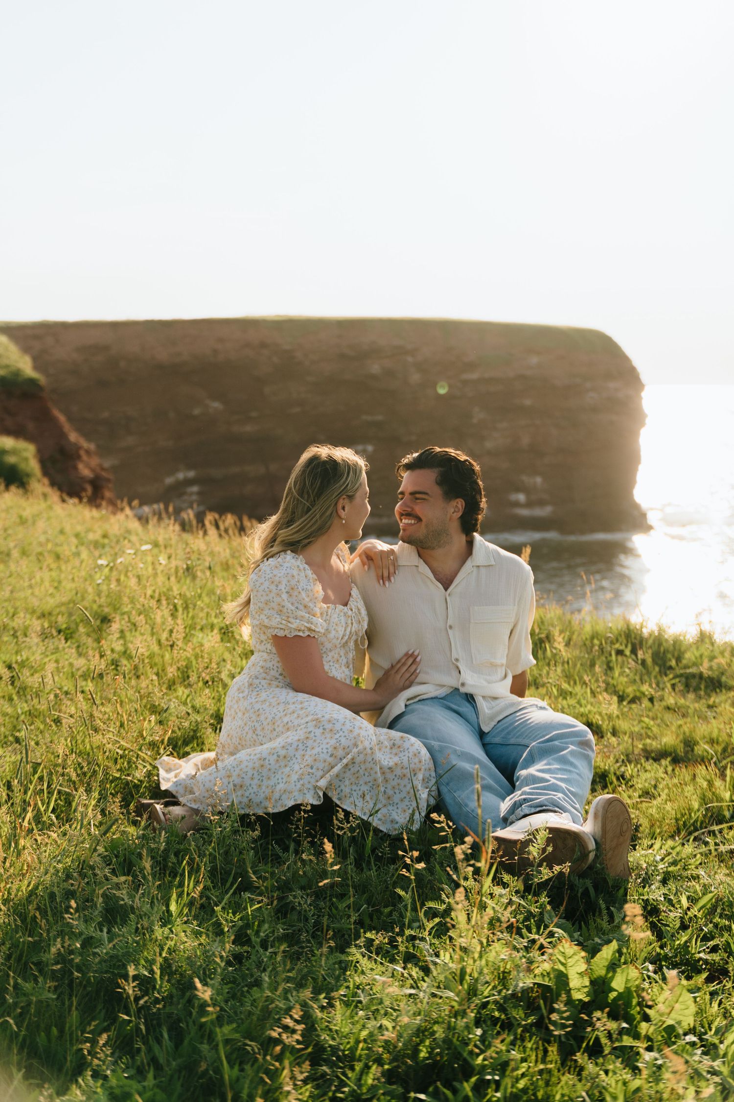 Couple sitting together on grassy clifftop overlooking the ocean at sunset.