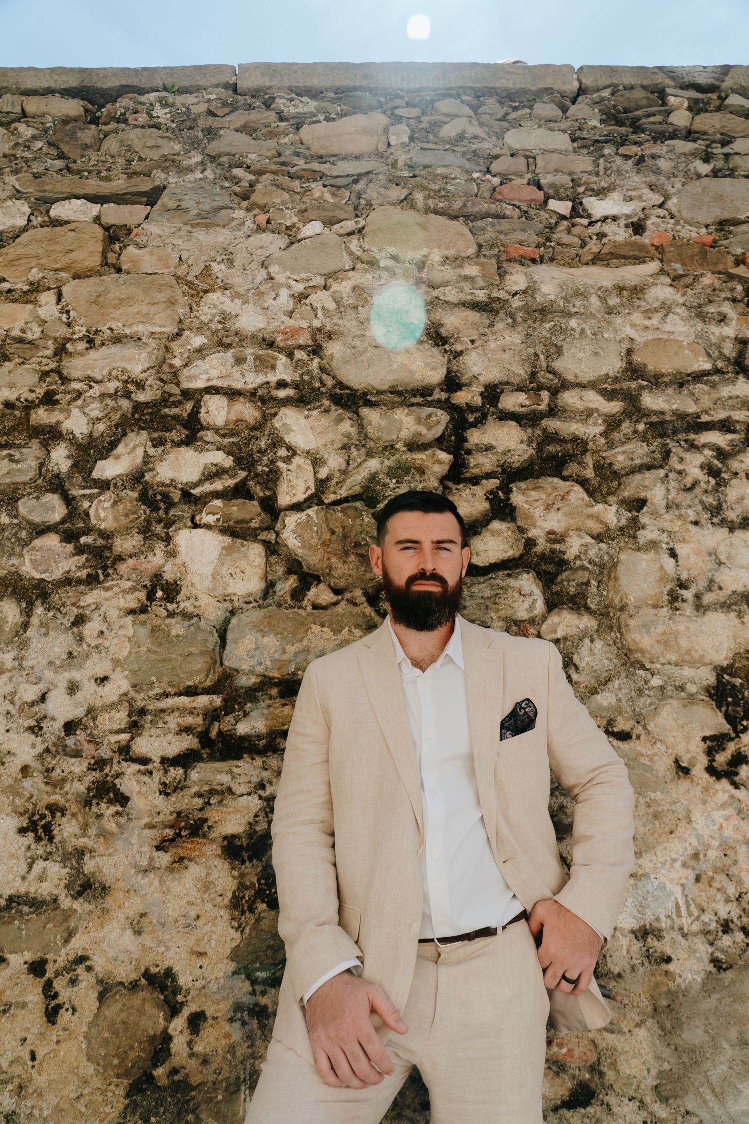 A groom in beige suit poses against a rustic stone wall background in Italian alley