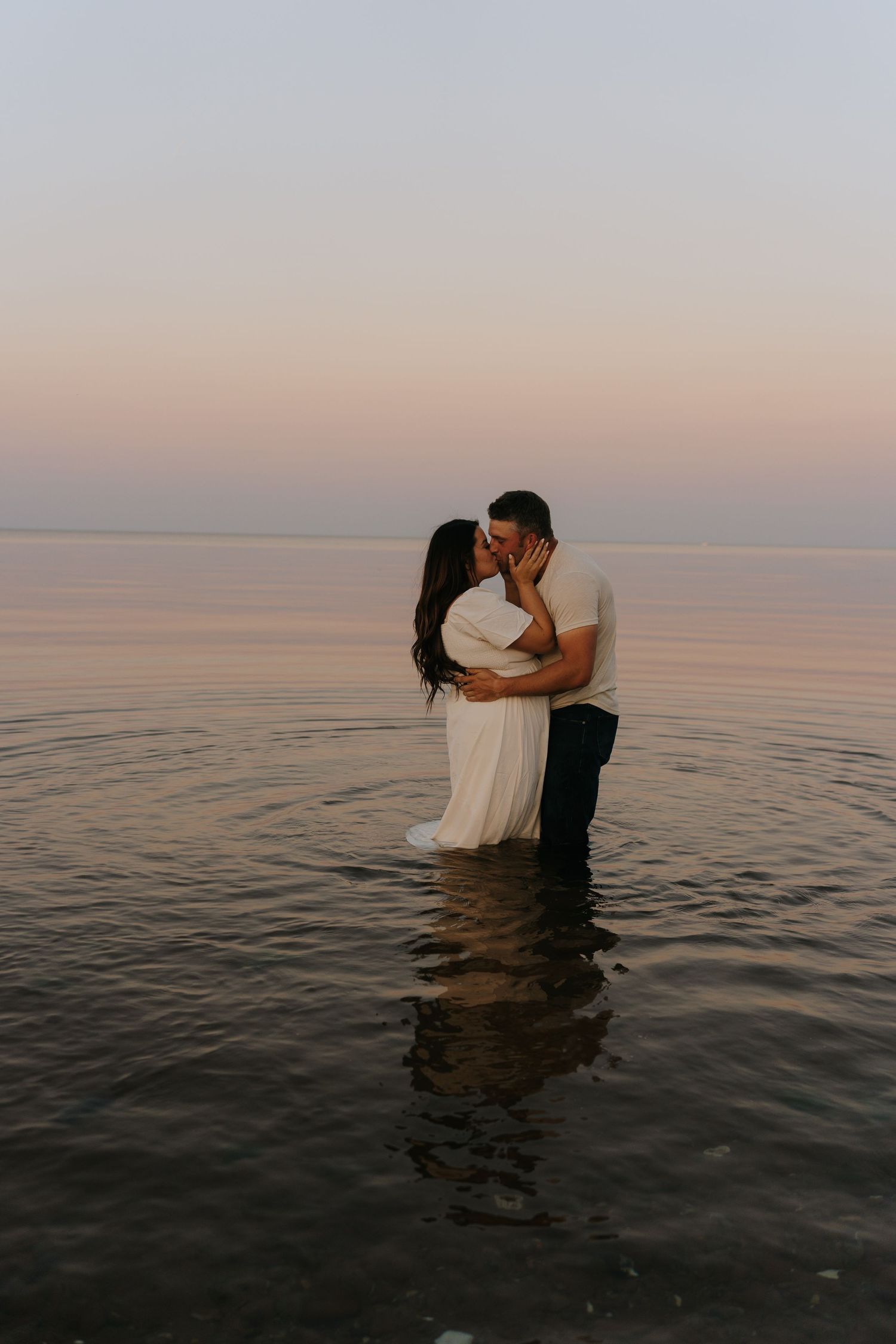 A couple shares a romantic moment while standing in calm waters at sunset.