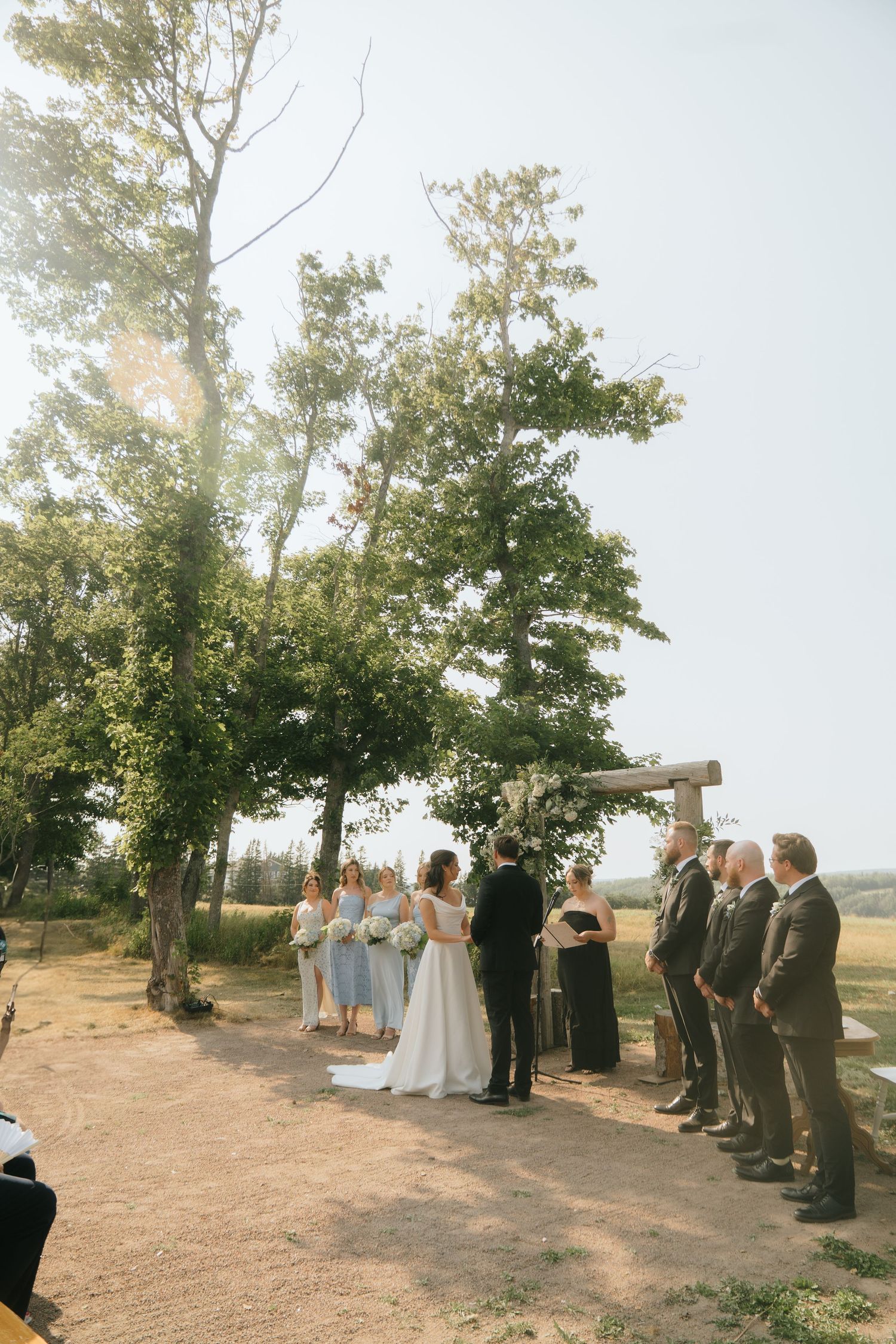An outdoor wedding ceremony under tall trees with guests lined up on both sides.