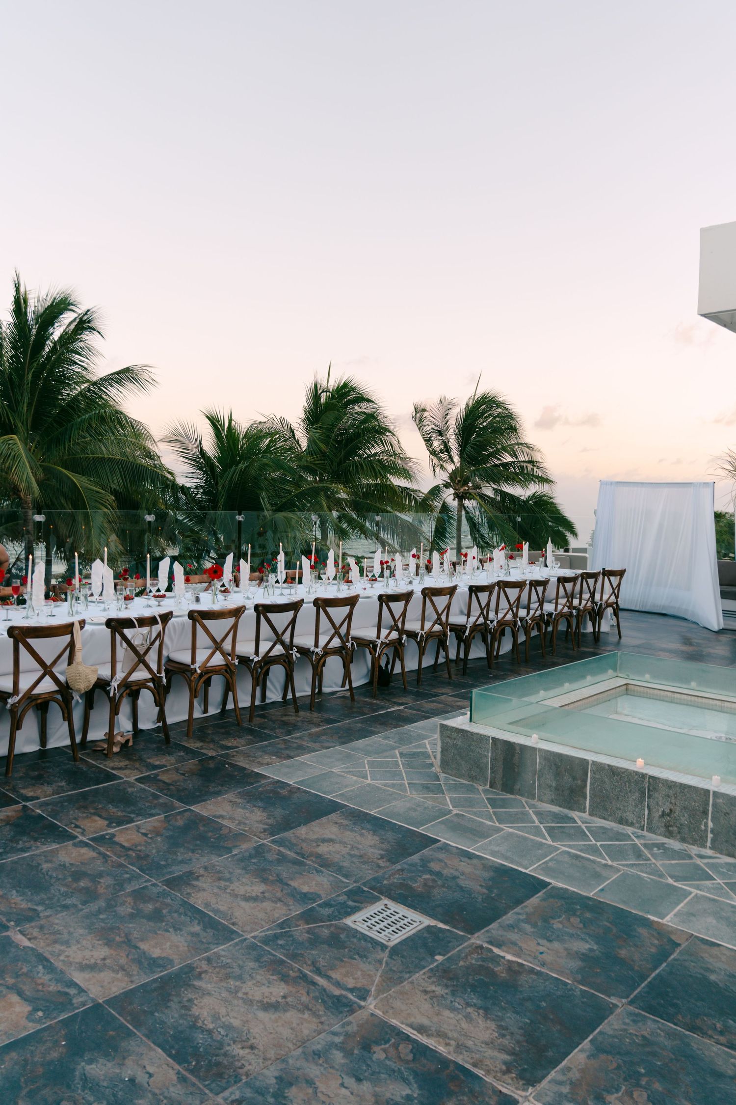 Elegant outdoor wedding reception with long dining table, wooden chairs, white linens, and tropical palm trees at sunset.