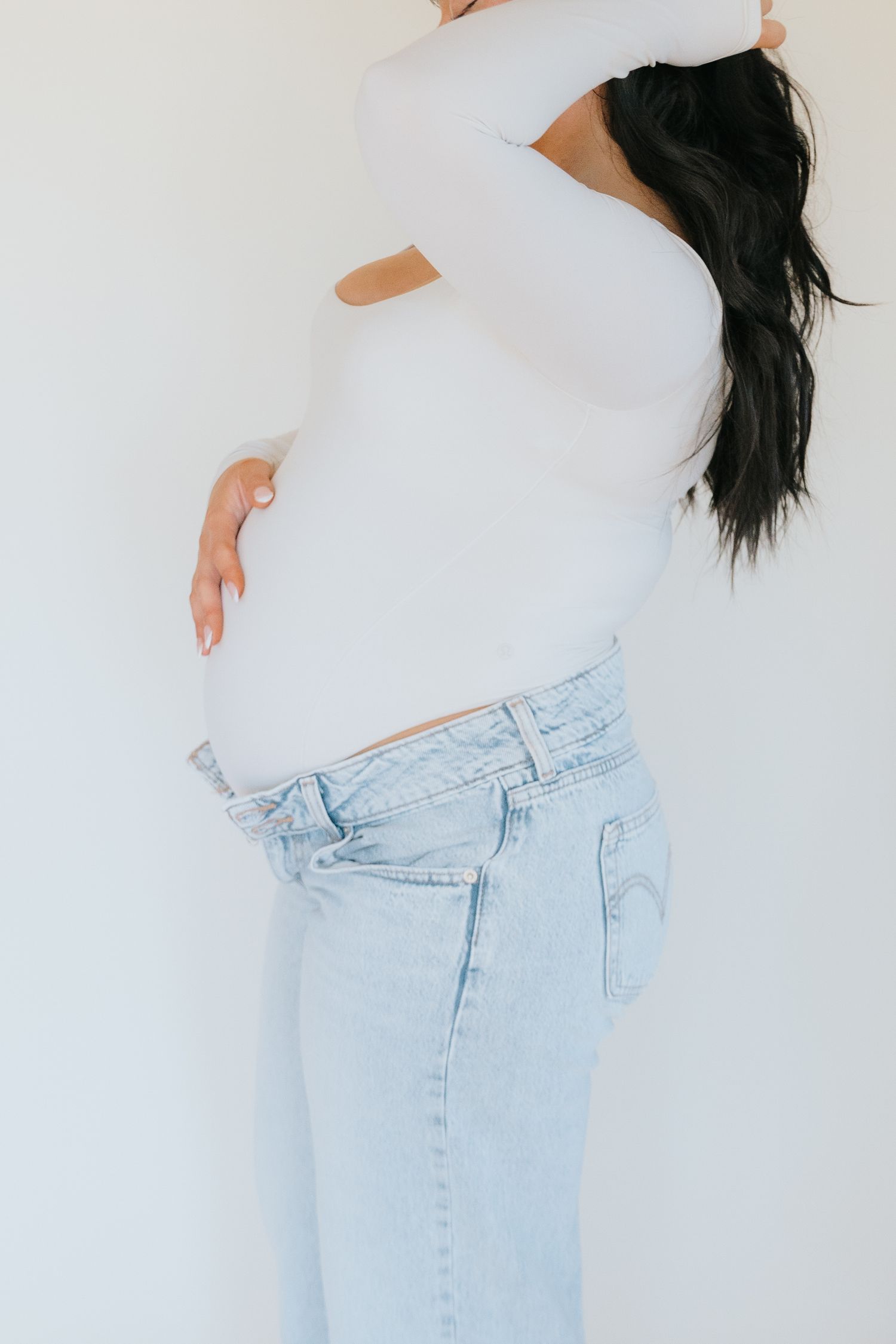 Pregnant woman in white top and light wash jeans cradling baby bump against white background.