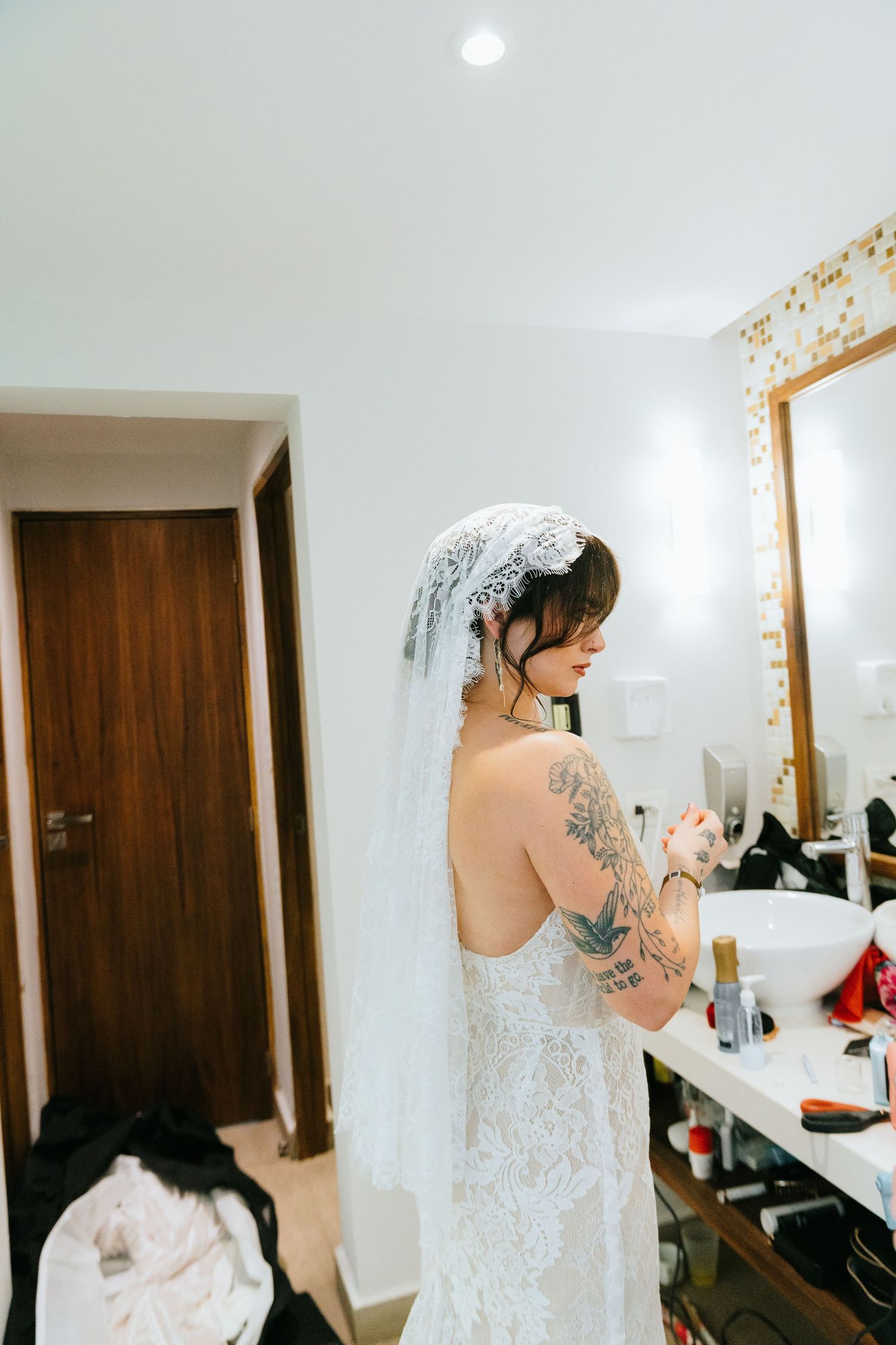 Tattooed bride in backless lace wedding dress and veil preparing at bathroom vanity mirror.