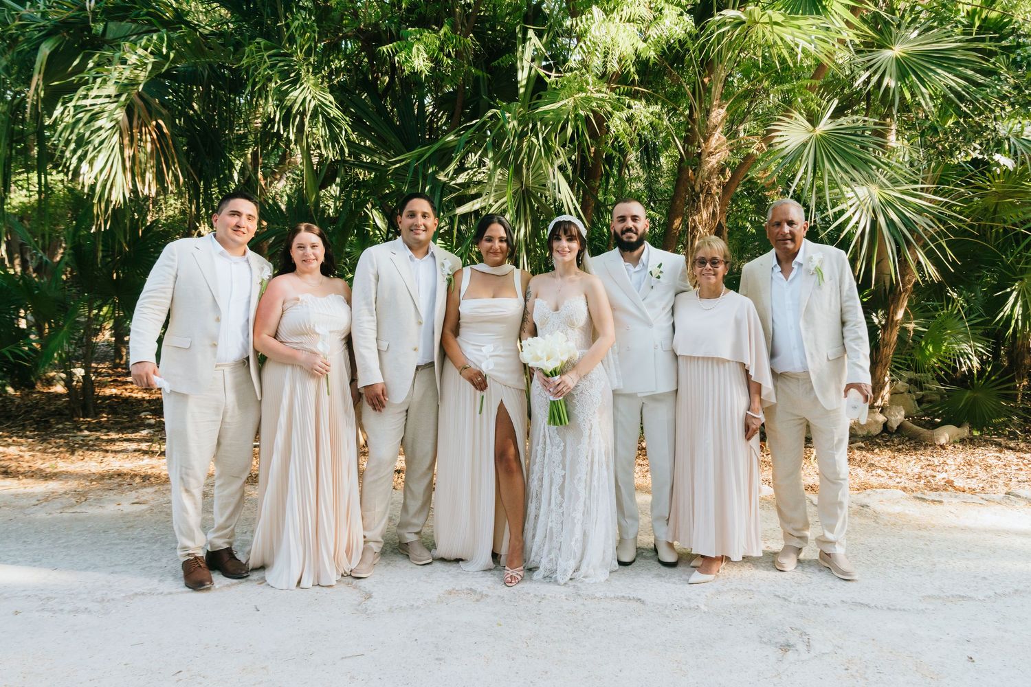 Wedding party poses in neutral attire among tropical palms at outdoor ceremony.