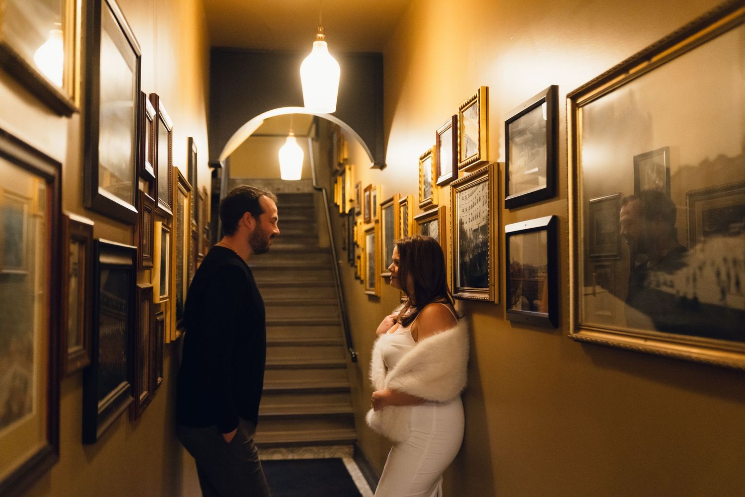 Couple sharing an intimate moment in a warmly lit hallway adorned with framed artwork and a staircase backdrop.
