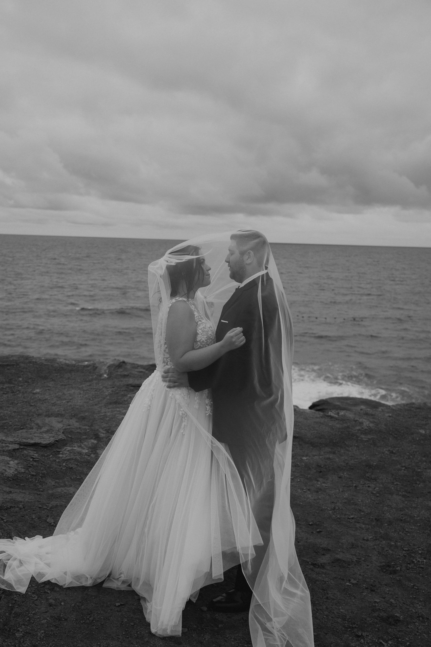 A romantic black and white wedding photo shows a couple embracing on a dramatic rocky coastline under moody clouds.