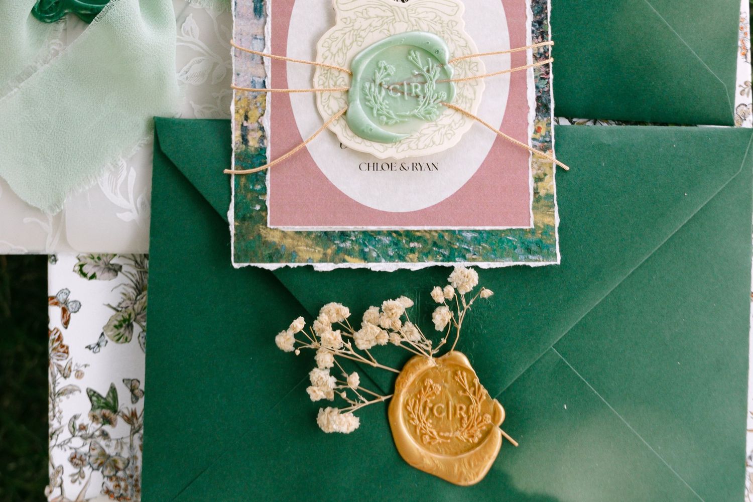 Elegant wedding stationery suite with green envelopes, gold wax seals, and dried flowers on a green background.