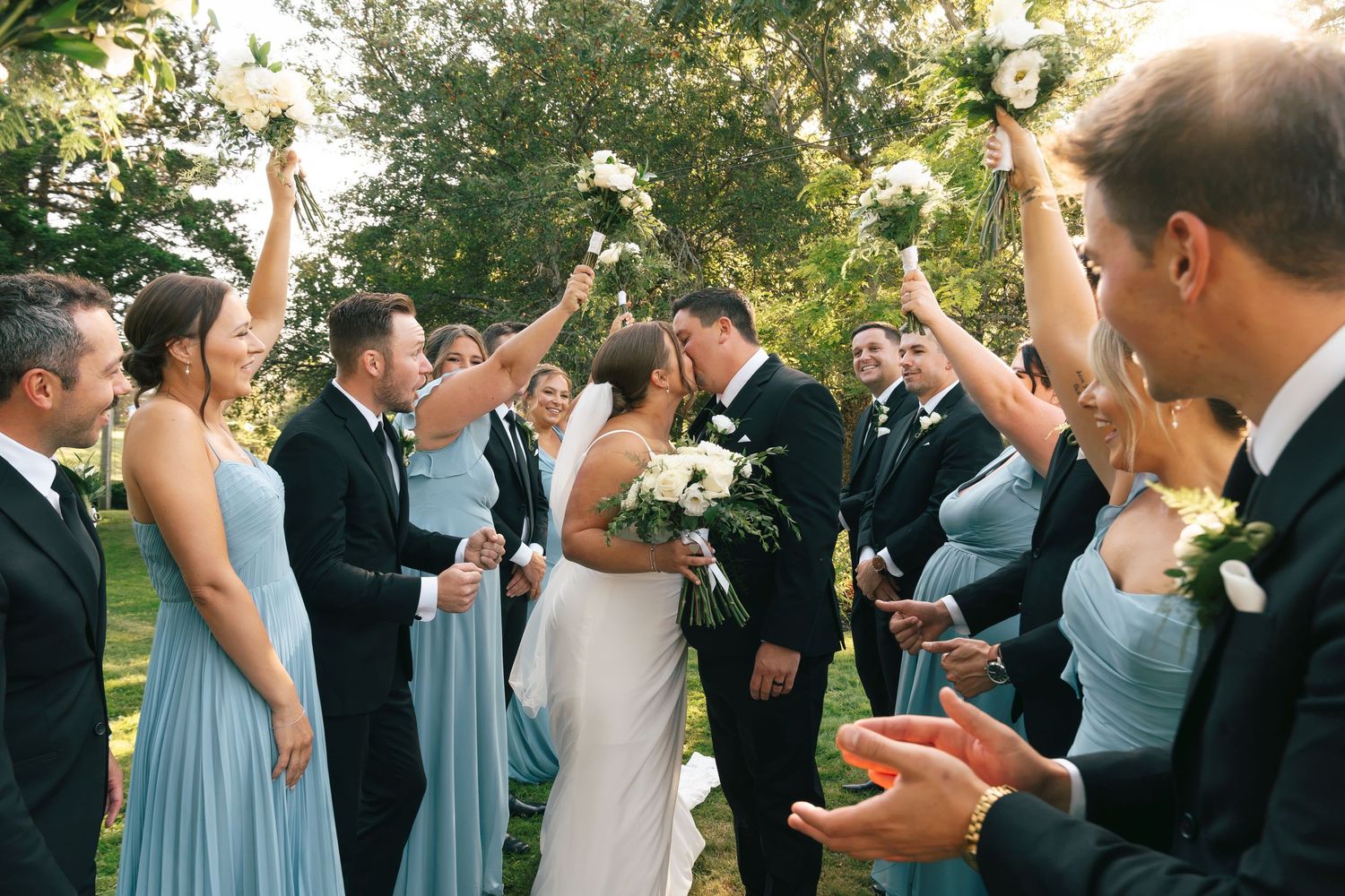 Bride and groom share a kiss surrounded by cheering wedding party in blue bridesmaid dresses outdoors.
