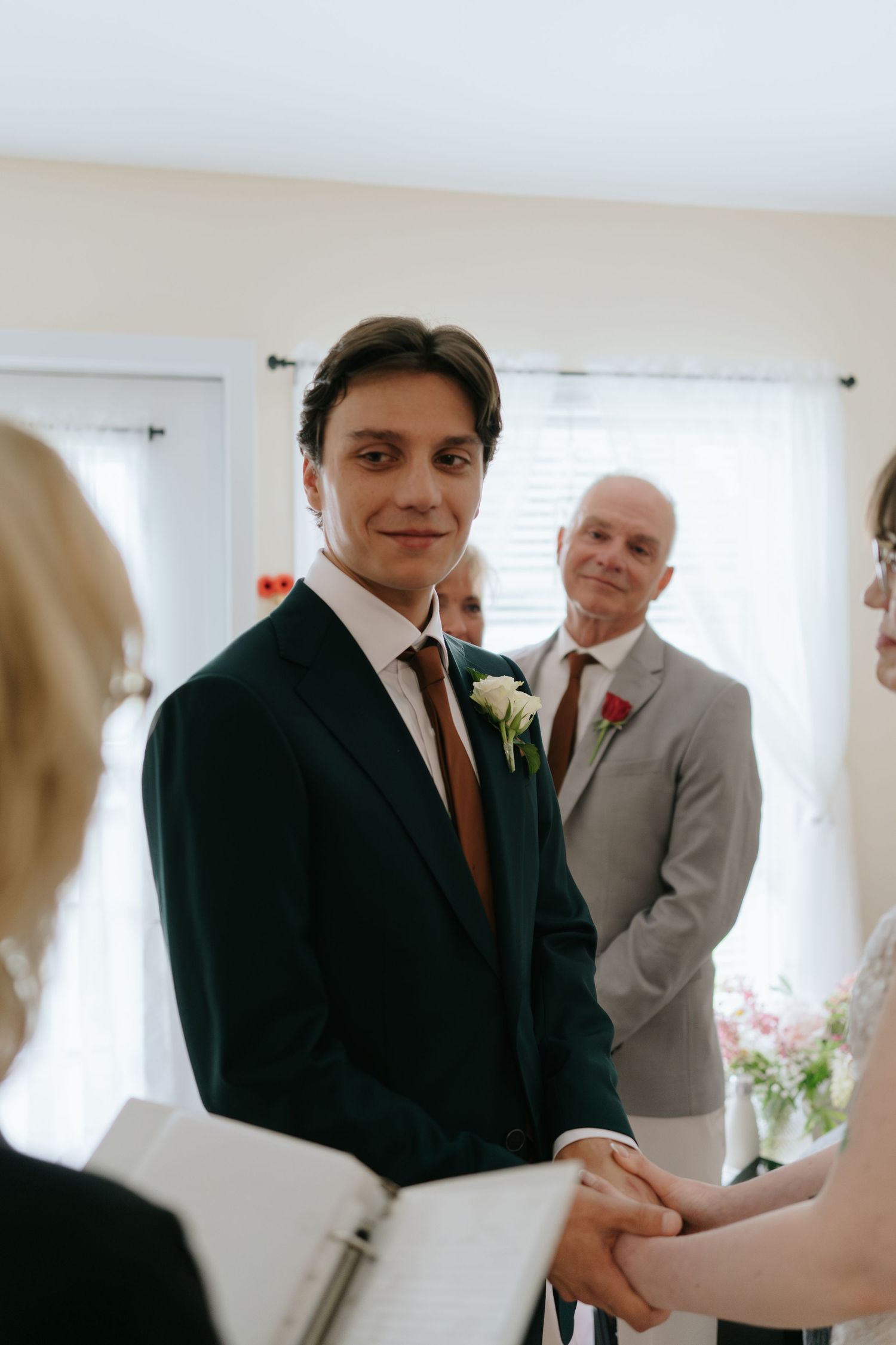 A wedding ceremony takes place indoors with attendees gathered around a couple exchanging vows.
