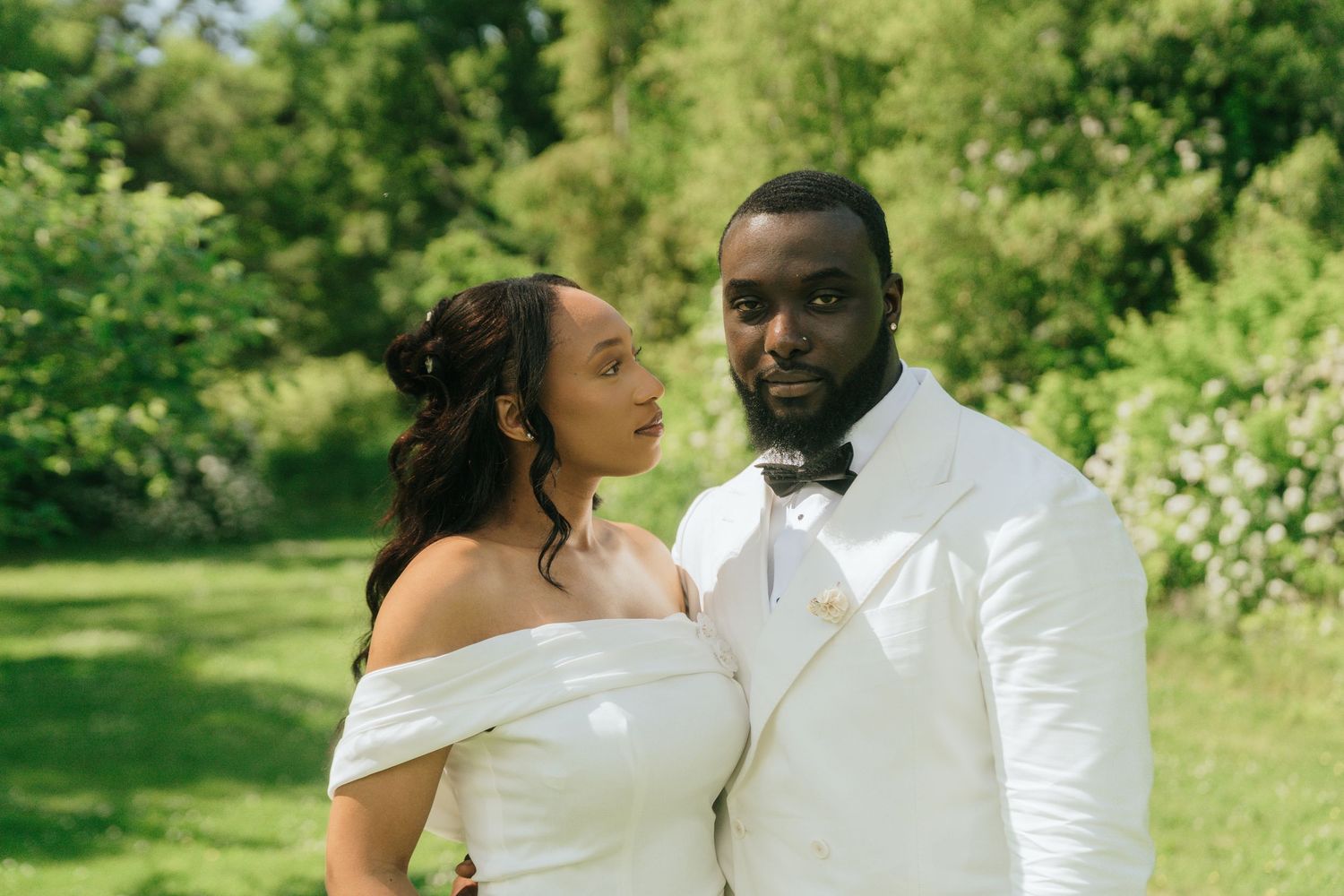 A couple in elegant white wedding attire poses together in a sunny garden setting.