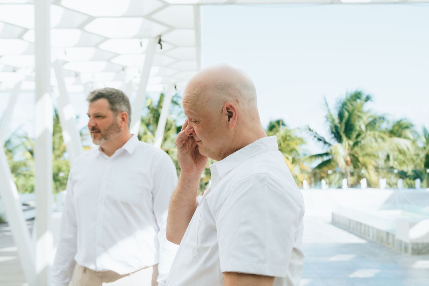 Two elderly men in white shirts stand on a tropical outdoor terrace with palm trees in the background.