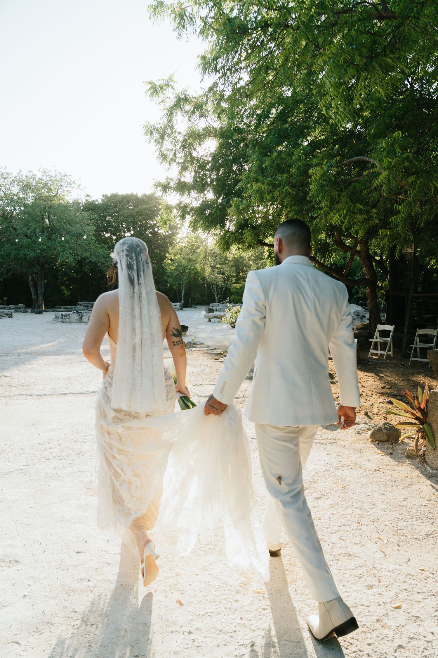 Bride and groom walking hand in hand on sandy path surrounded by lush green trees during outdoor wedding ceremony.