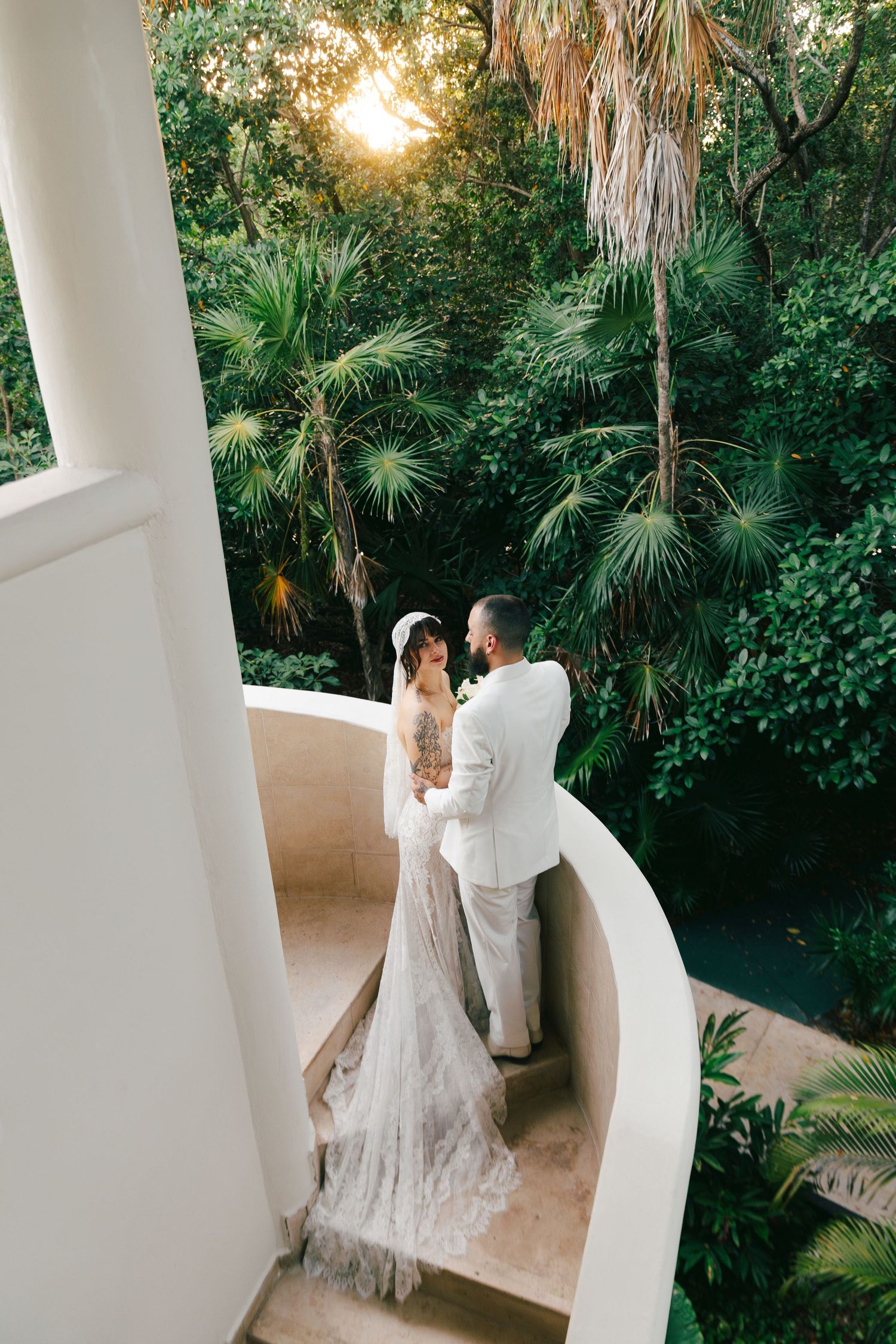 Bride and groom share a romantic moment on a curved balcony surrounded by lush tropical greenery at sunset.