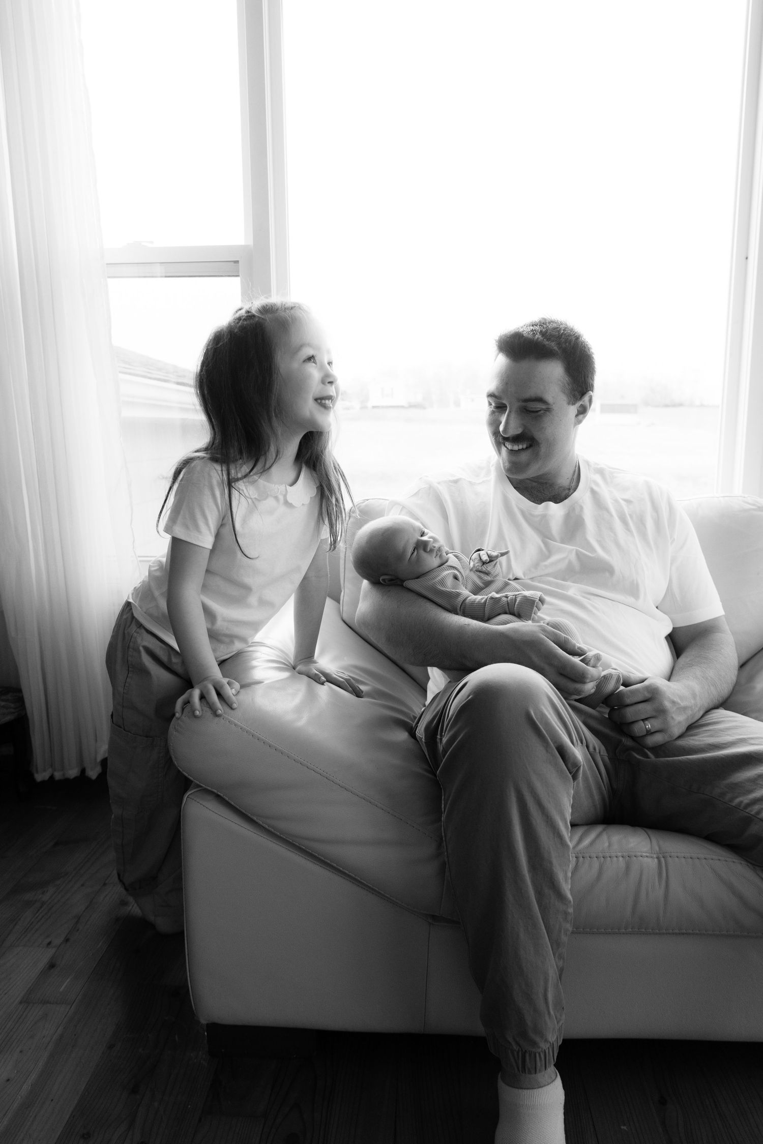 Black and white photo of a smiling father sitting on a couch holding a newborn baby with his young daughter beside him.