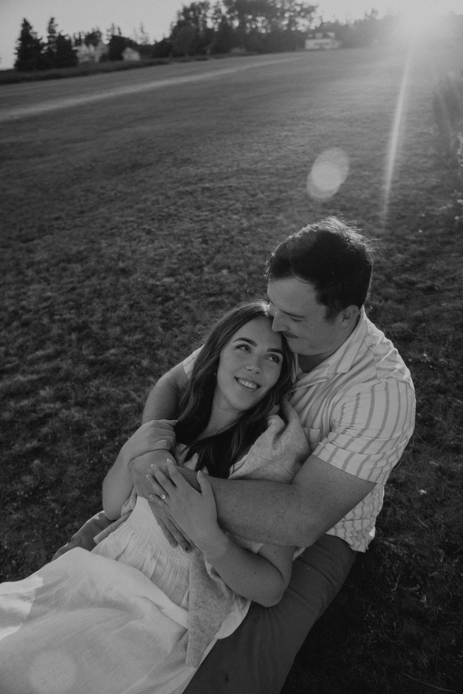 Black and white photo of a couple sitting in a field at sunset, woman smiling up as man embraces her tenderly.