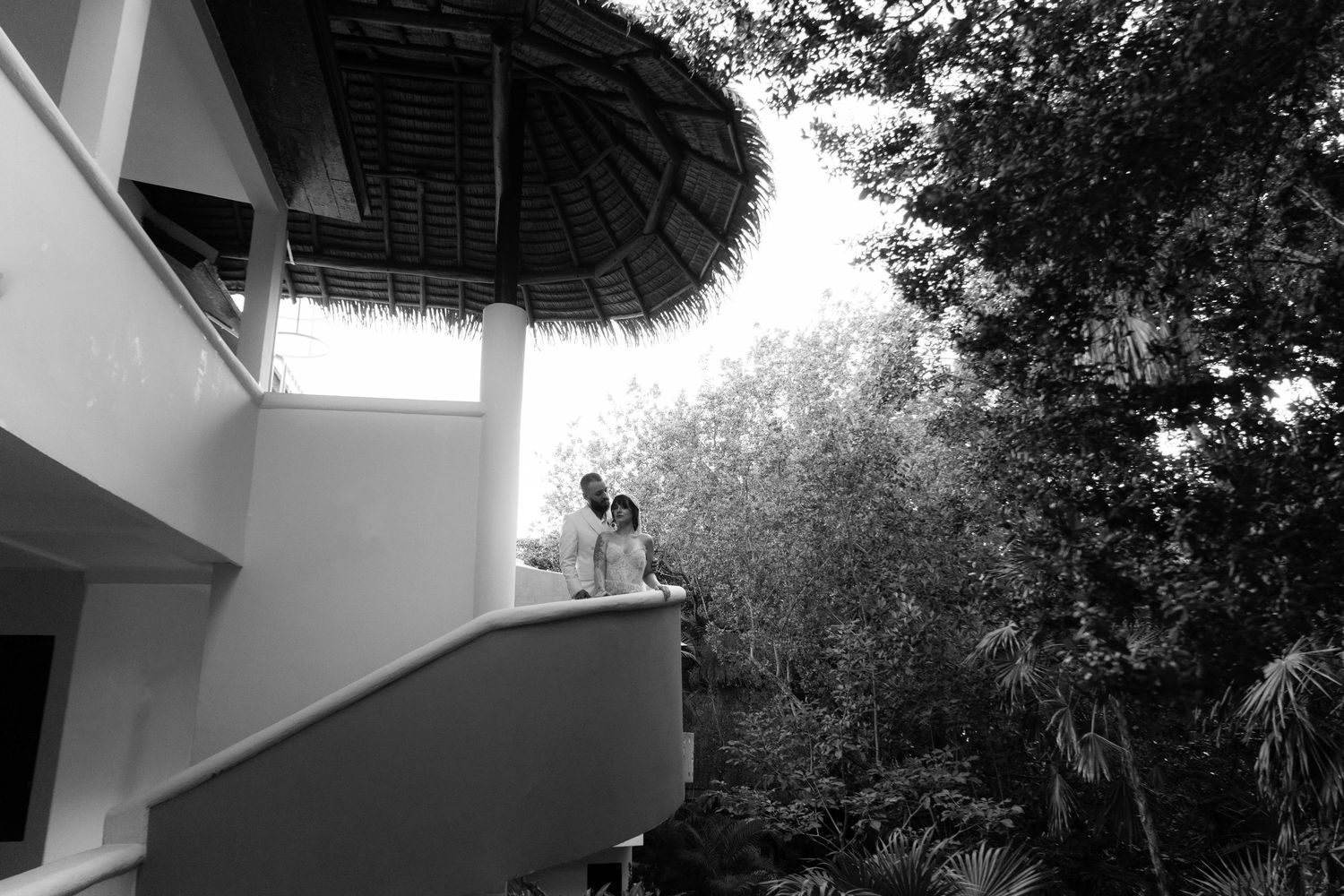 Black and white photo of a couple embracing on a balcony beneath a thatched palapa roof surrounded by lush tropical trees  Playa Del Carmen