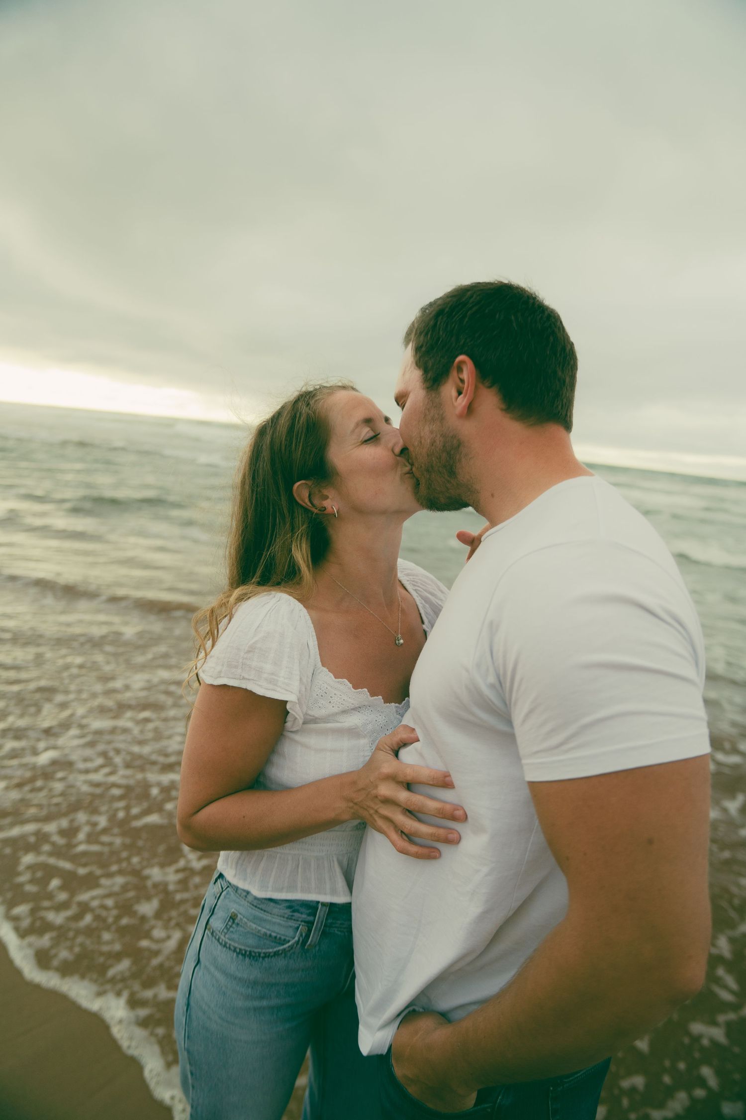 A couple shares a romantic kiss on a beach at sunset, both wearing white tops and jeans with ocean waves behind them.