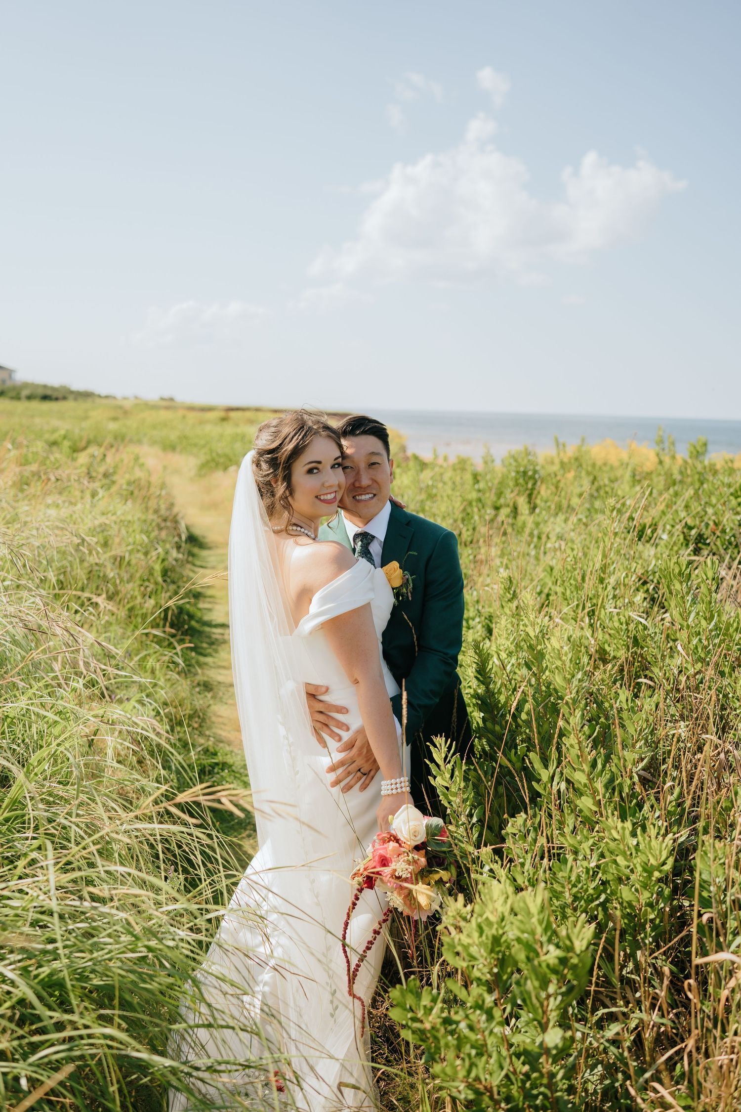 Wedding couple embraces in tall grass with ocean views and bright blue sky.