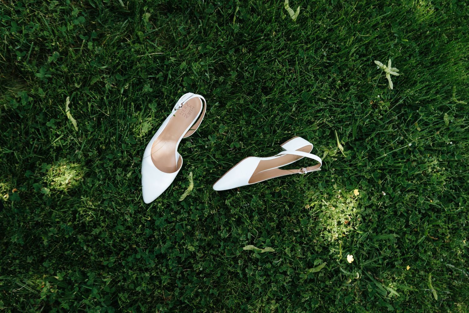 White bridal shoes lying on lush green grass in natural sunlight.