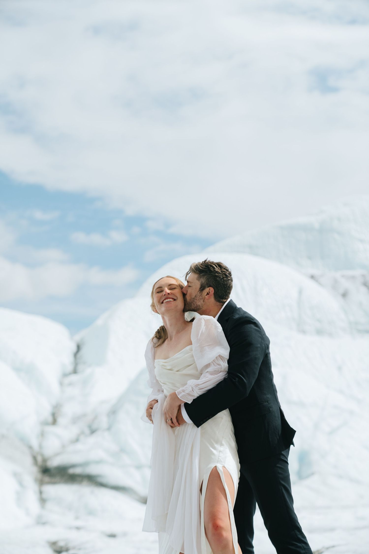 Couple in wedding attire embracing on a snow-covered glacier with mountain backdrop in Alaska