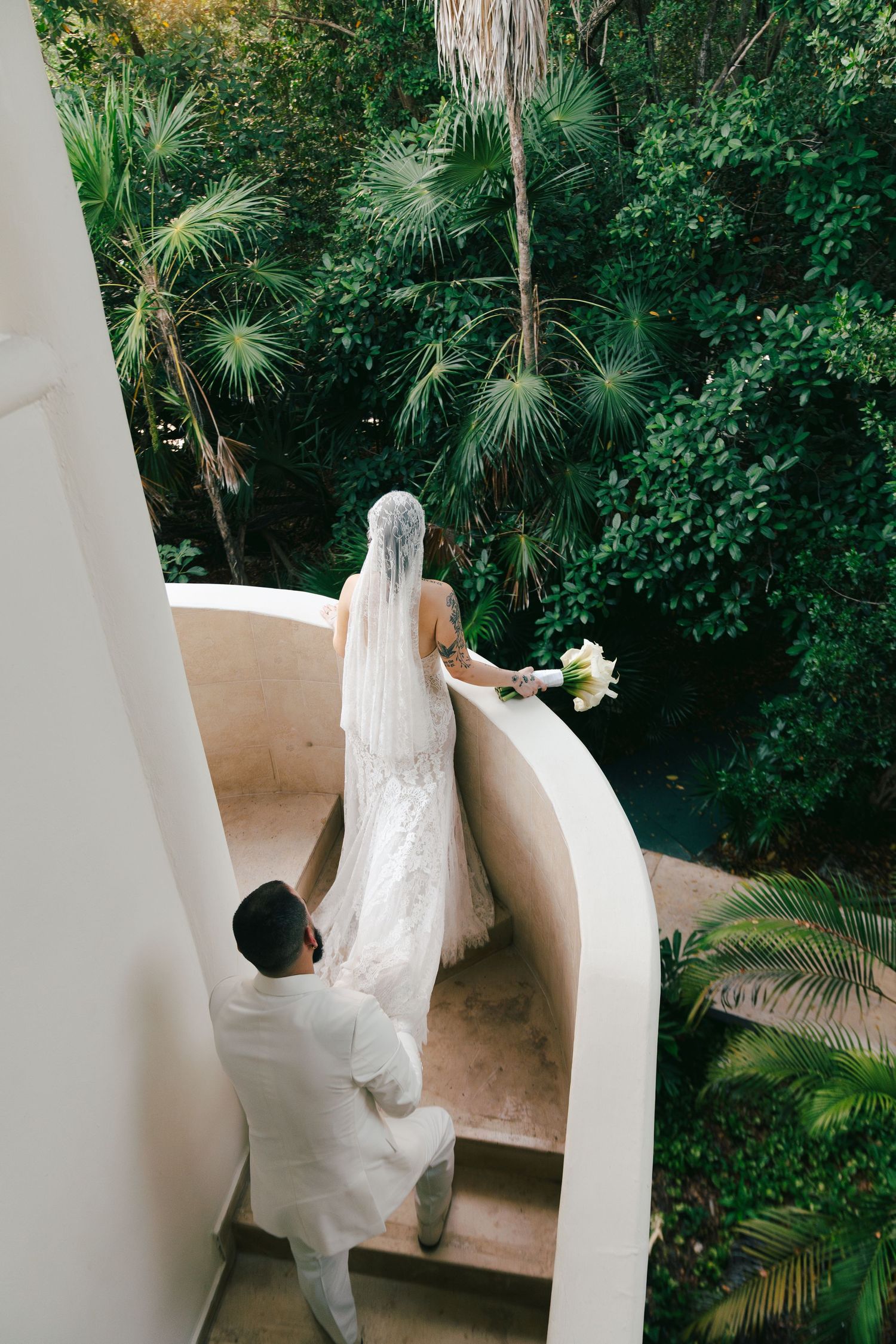 Bride in white gown and veil with groom ascending curved staircase overlooking lush tropical garden.