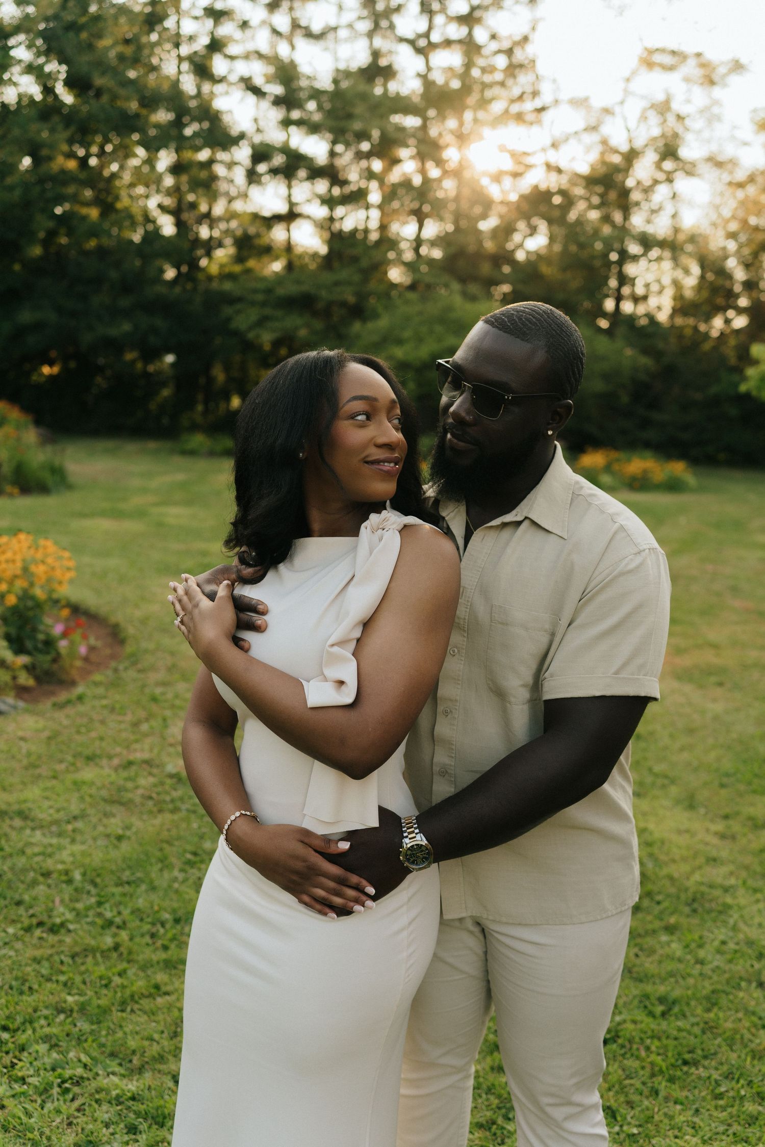 Couple in light-colored outfits sharing intimate moments in a garden setting at sunset.