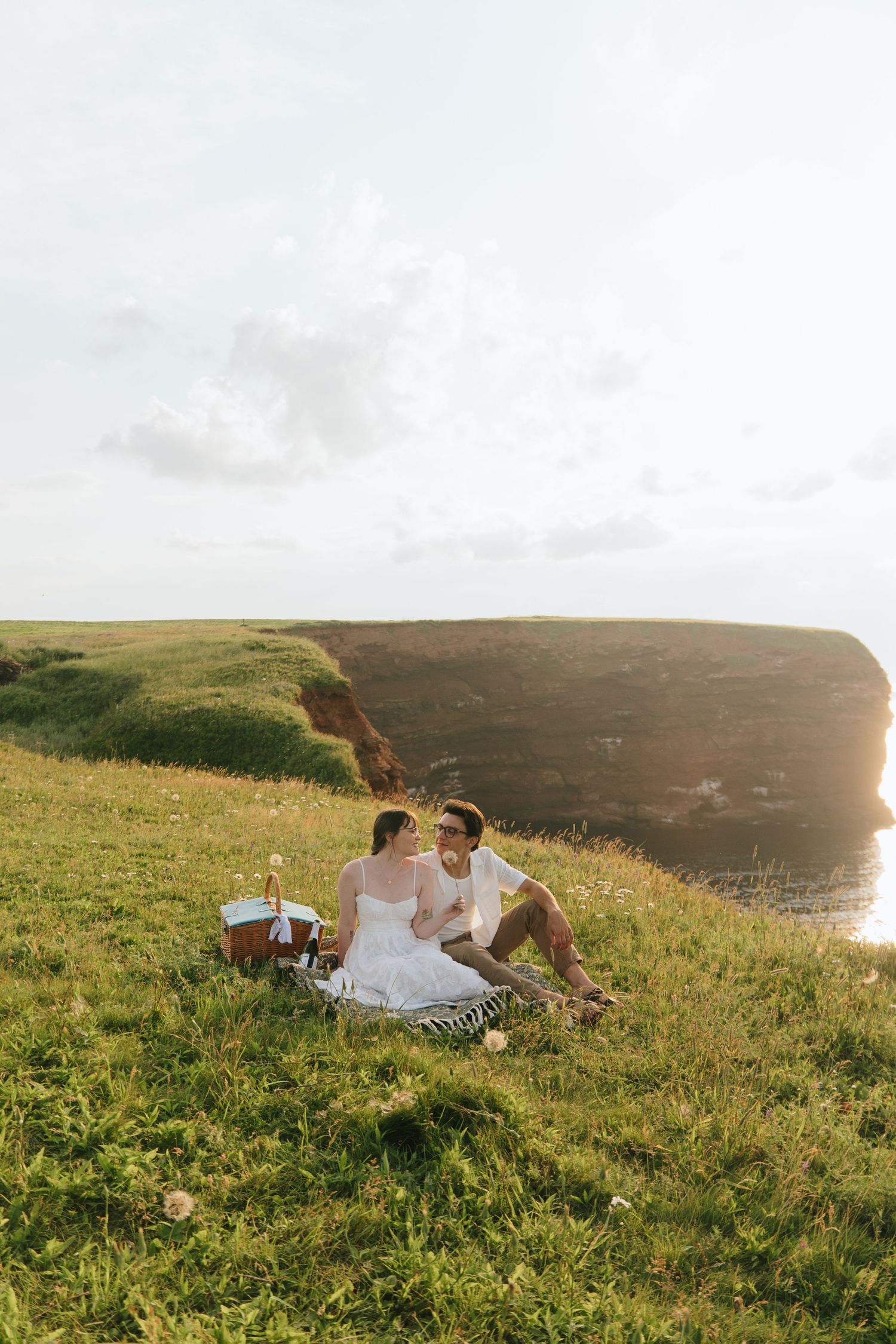 Couple enjoying a romantic sunset picnic on a grassy coastal hillside overlooking the ocean.