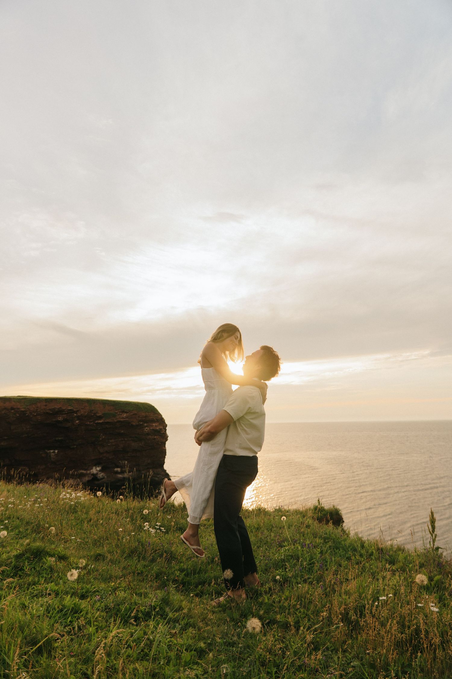 A couple embraces on a scenic coastal cliff at sunset with wildflowers in the foreground.
