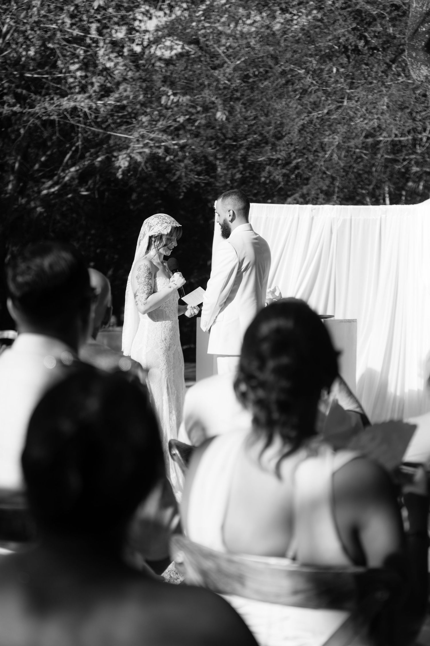 Black and white photo of a bride and groom exchanging vows outdoors, with guests watching the intimate wedding ceremony.