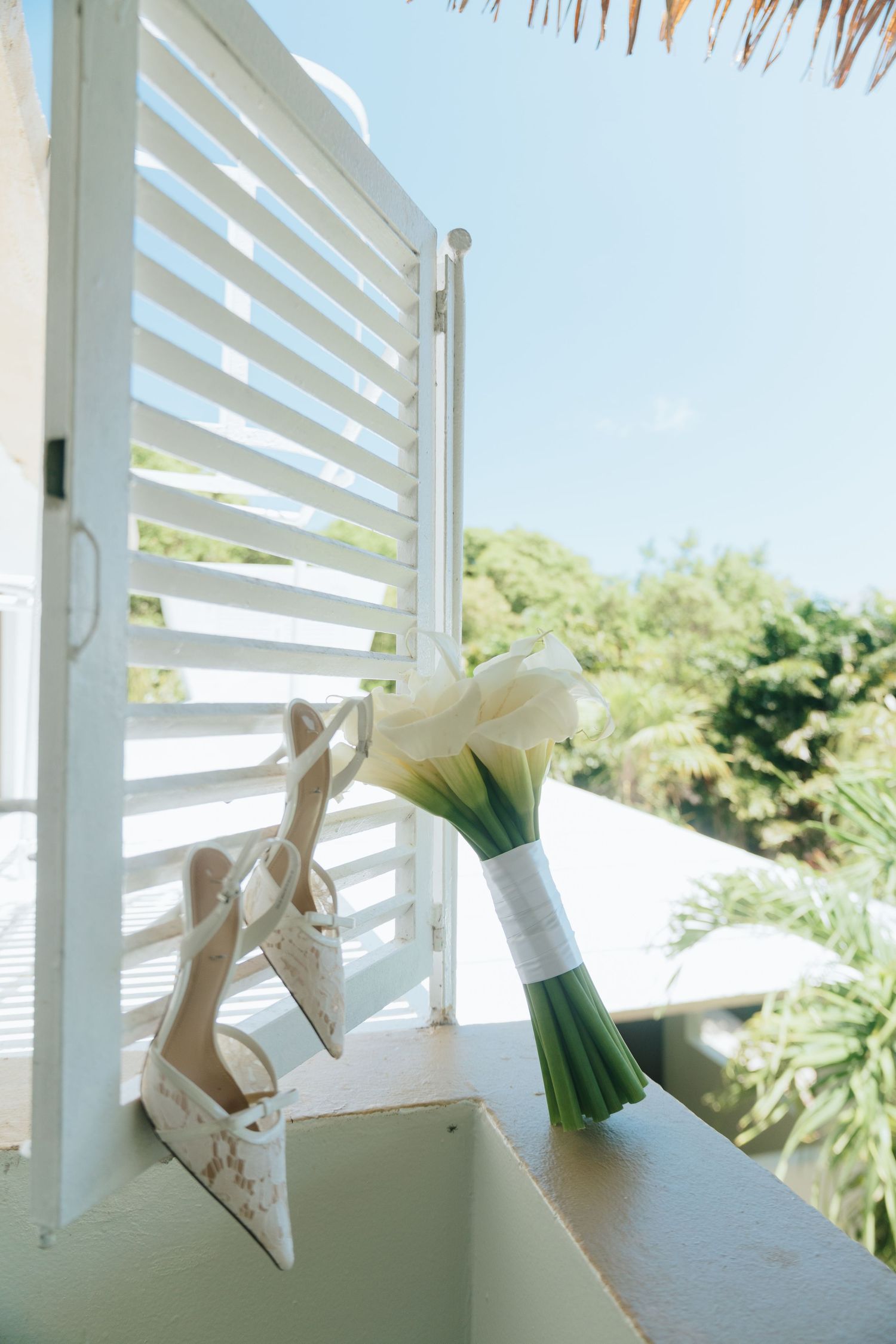 White calla lily bridal bouquet and embroidered heels displayed on a sunlit balcony railing with tropical greenery backdrop.