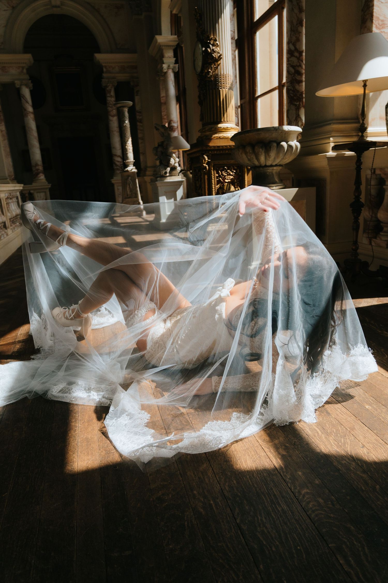 Artistic shot of a bride lying on hardwood floor draped in a sheer lace veil in a sunlit ornate hall.