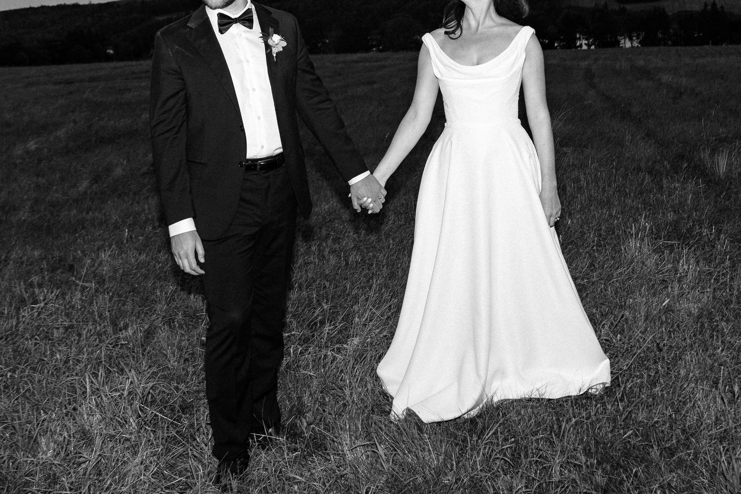 Black and white photo of a couple holding hands while walking through a meadow in formal attire.