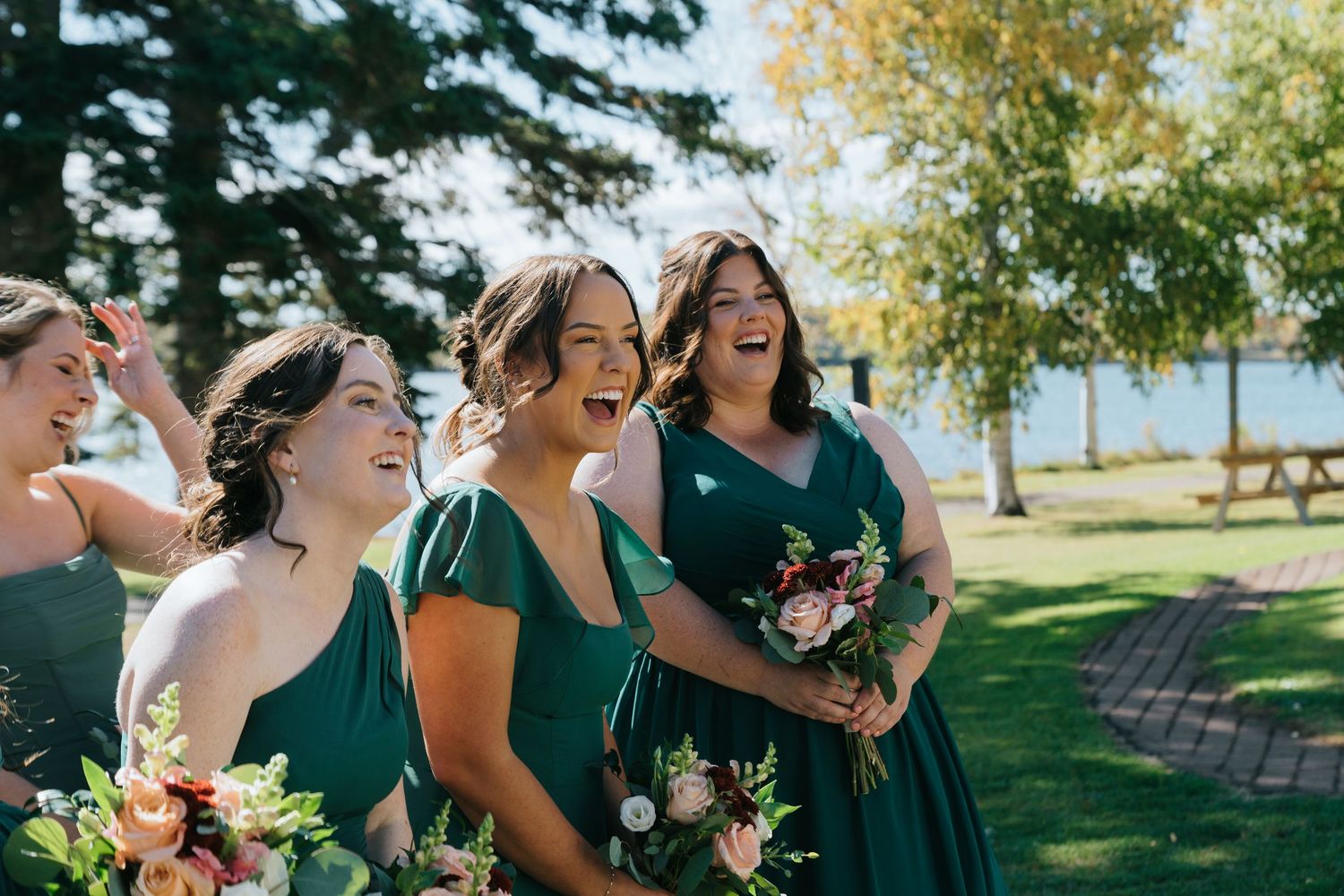 Bridesmaids in emerald green dresses laughing and holding floral bouquets outdoors near a lake on a sunny day.
