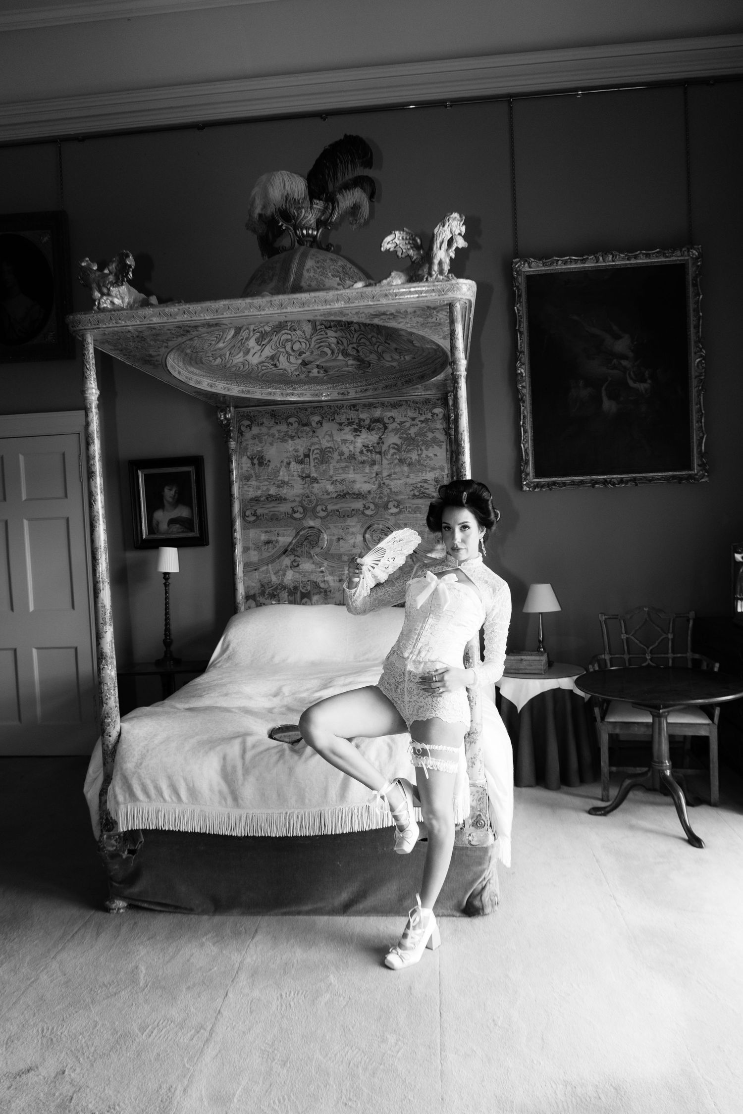 Black and white photo of a woman in lace lingerie posing by an ornate four-poster bed in a grand bedroom.