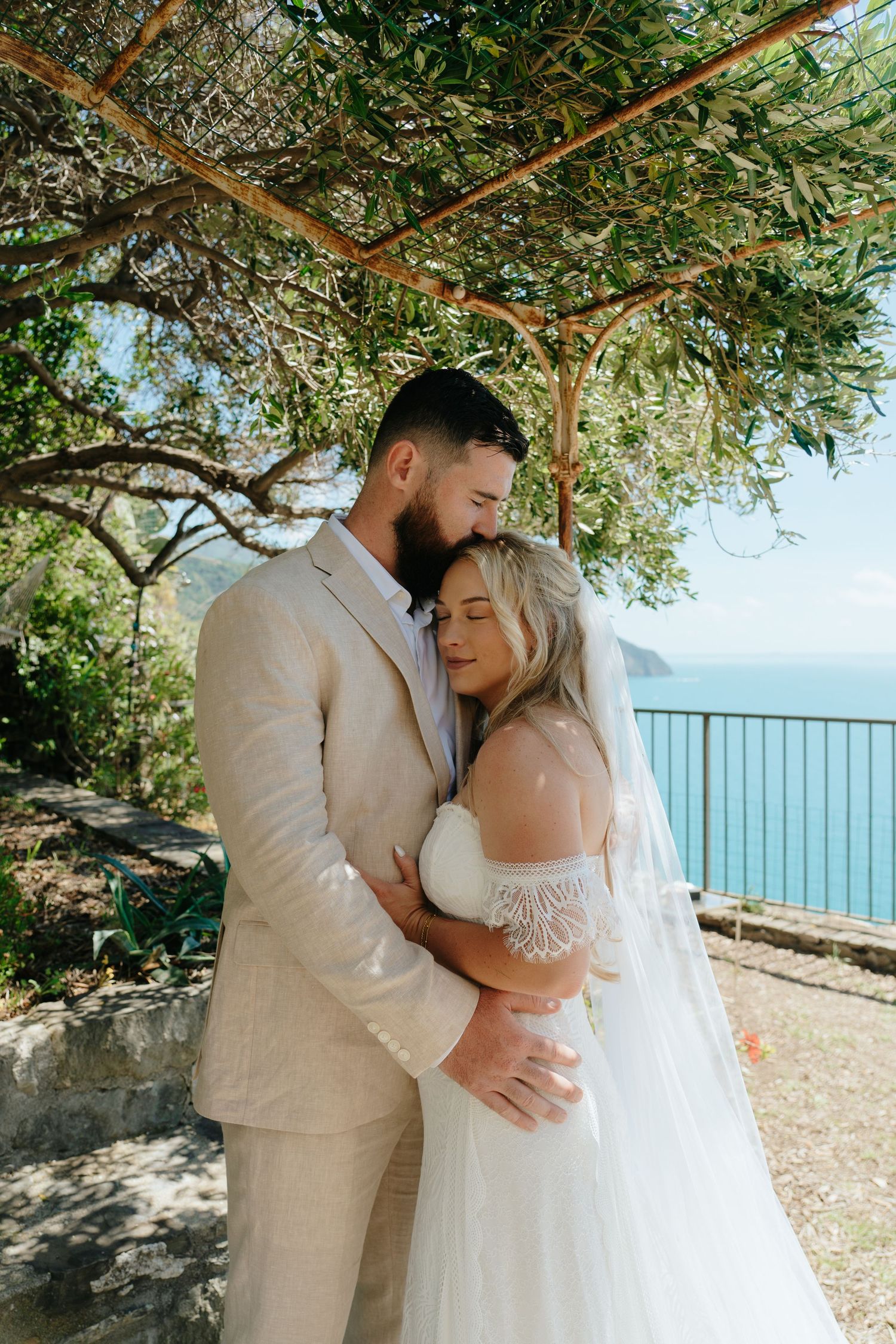 A romantic wedding couple embraces under an olive tree pergola overlooking a scenic coastal view on a sunny day.