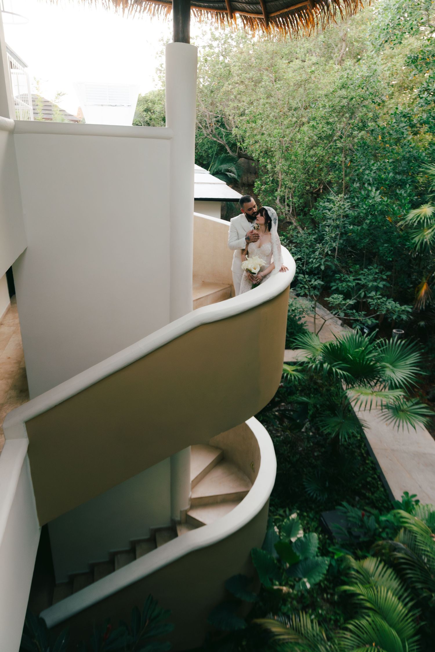 Couple embracing on a curved spiral staircase surrounded by lush tropical greenery at a resort.