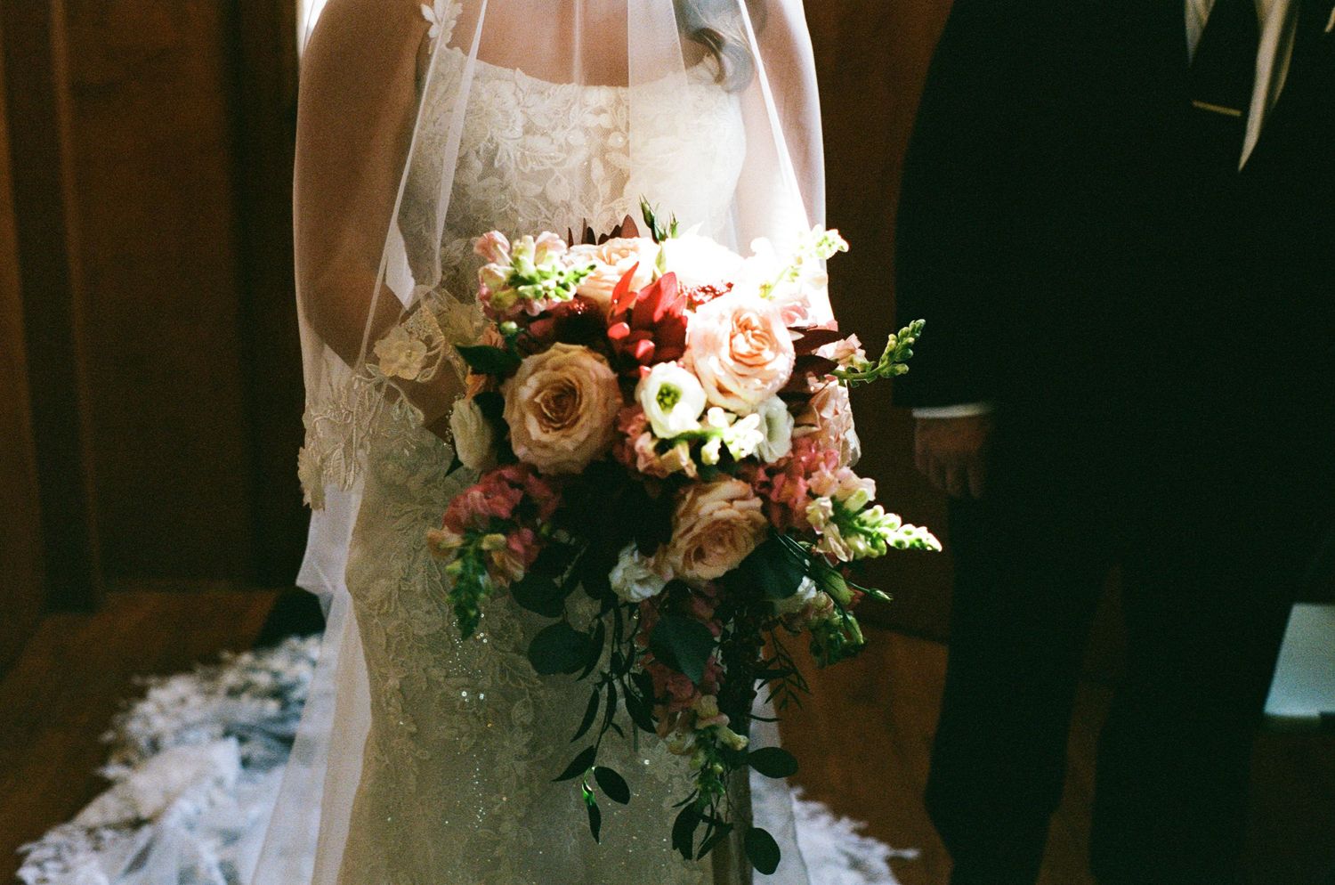 A bride in a white lace wedding dress holds a romantic bouquet of pink and white flowers in natural lighting.