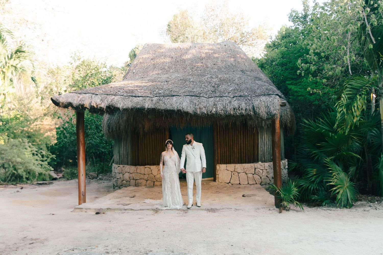 Bride and groom stand together under a thatched palapa structure surrounded by tropical jungle at their destination wedding.