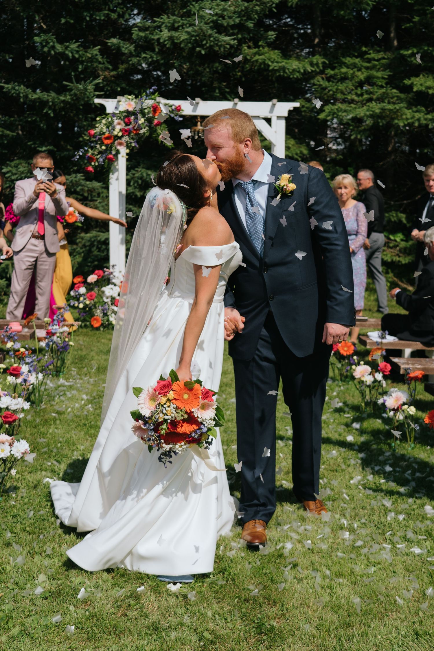Wedding ceremony guests toss flower petals as the couple walks through on a sunny garden path.