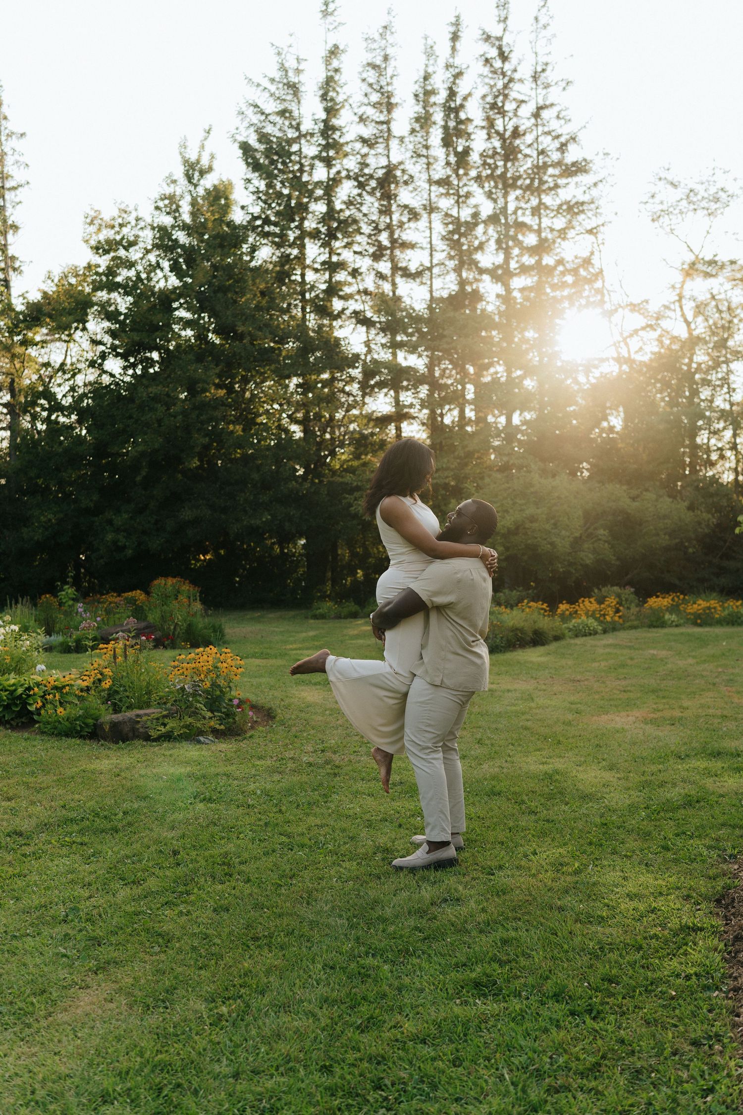 Couple embracing in a garden with evergreen trees and golden sunlight.
