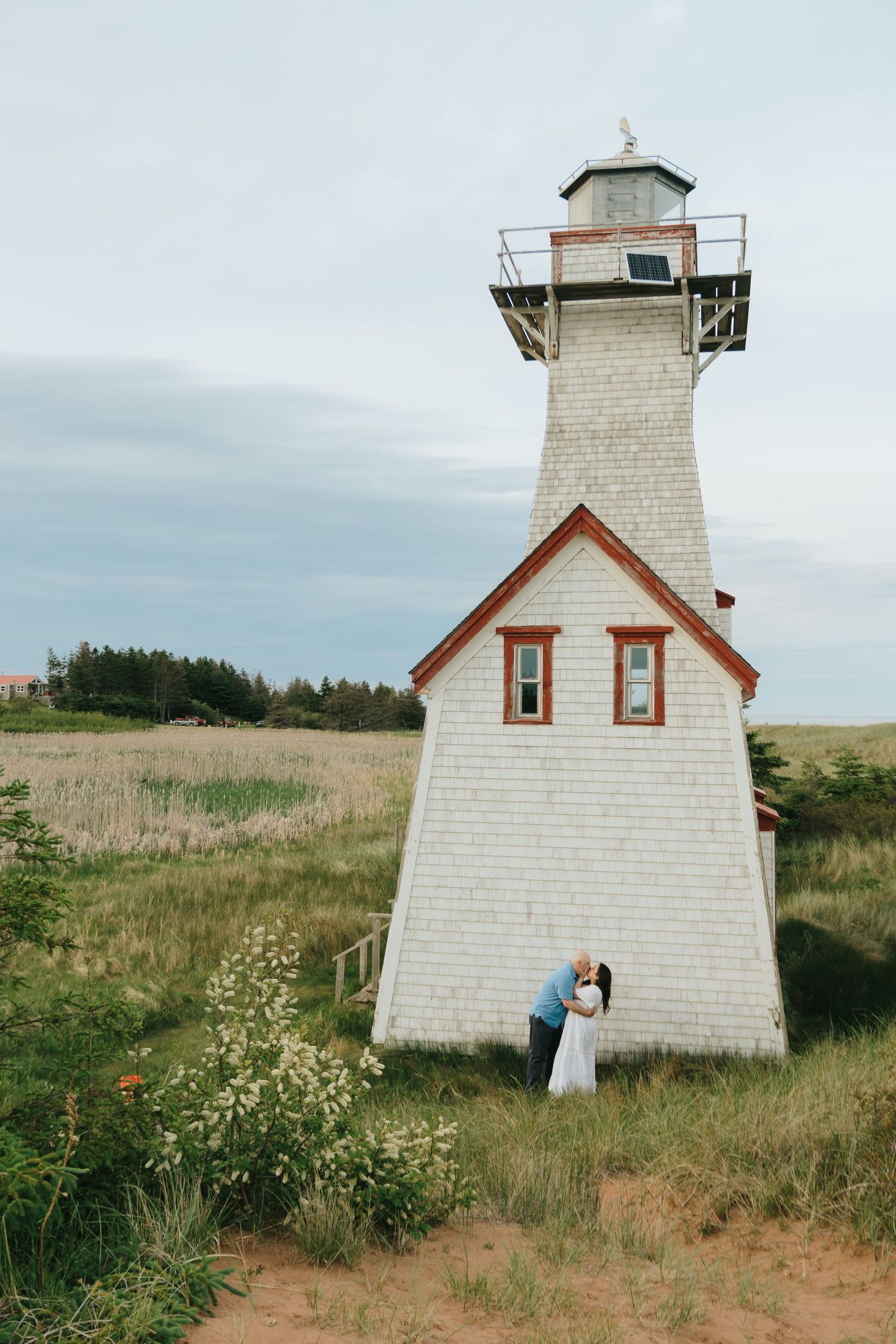 Couple kissing beside a white shingled lighthouse surrounded by golden fields and wildflowers under a cloudy sky.