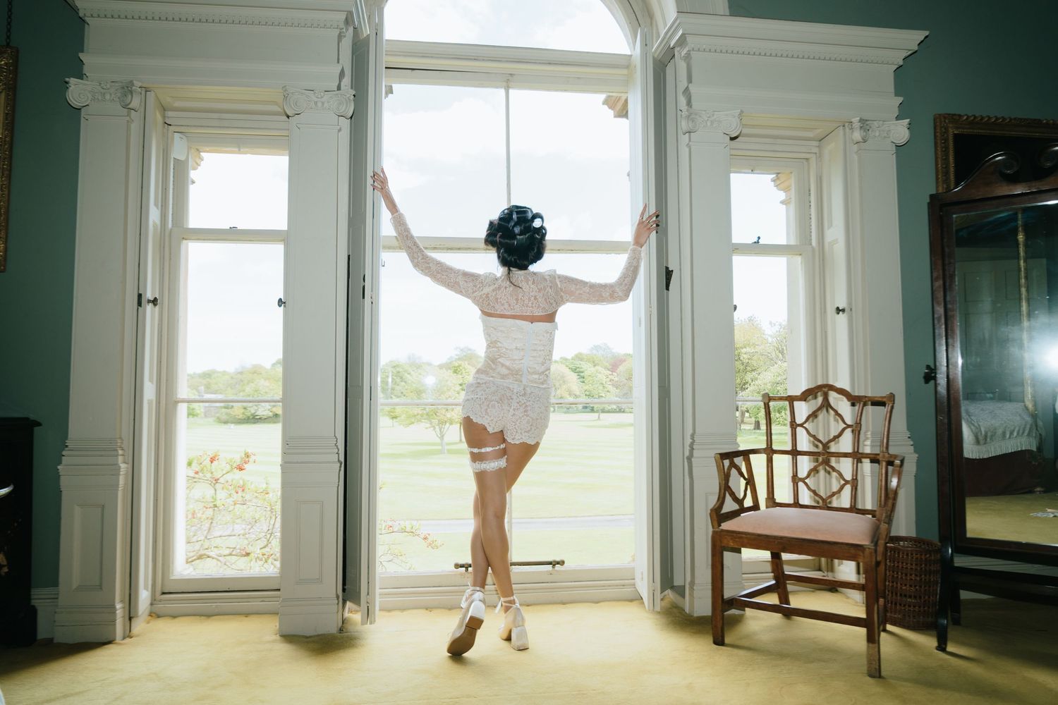 Woman in white lingerie and heels stands at tall elegant window overlooking green countryside in a grand room.
