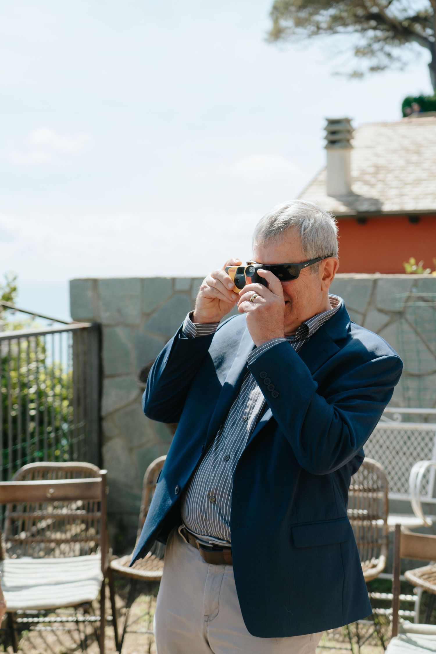 A photographer in a navy blazer takes pictures at an outdoor venue with mountain views in the background.