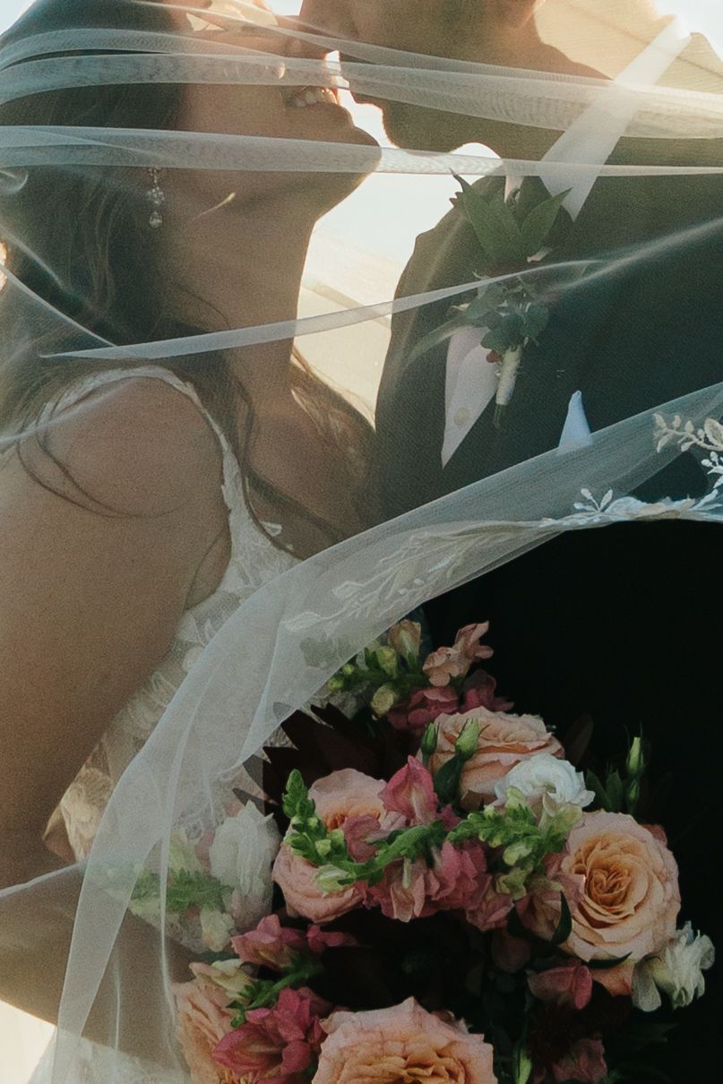 Bride and groom kissing under a flowing veil, holding a bouquet of pink and peach roses in golden sunlight.