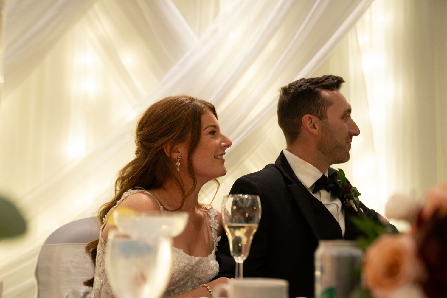 Bride and groom smiling at wedding reception table with champagne glasses, elegant draped white fabric backdrop.