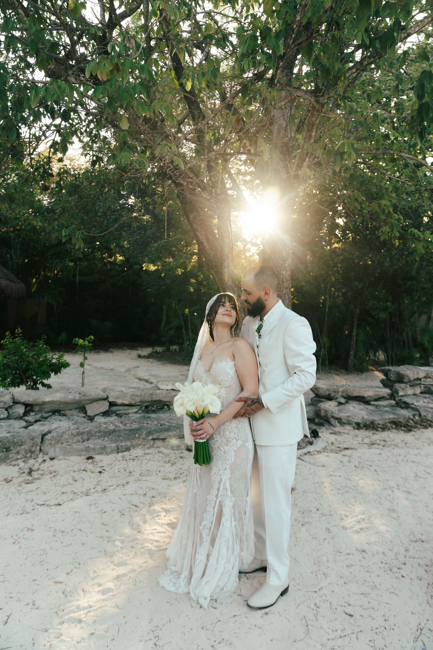 Couple embracing at sunset in tropical garden, bride in lace gown holding white bouquet with groom in white suit.