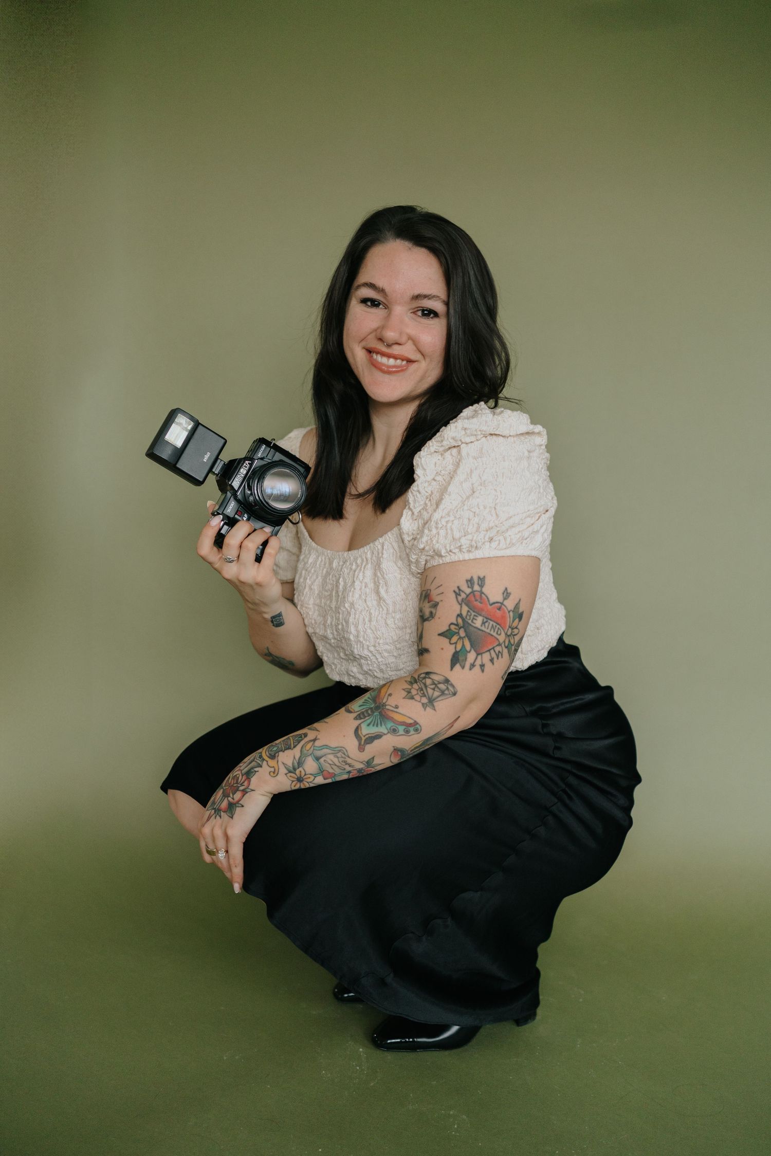 Professional photographer in white lace top and black skirt poses with camera against green backdrop.