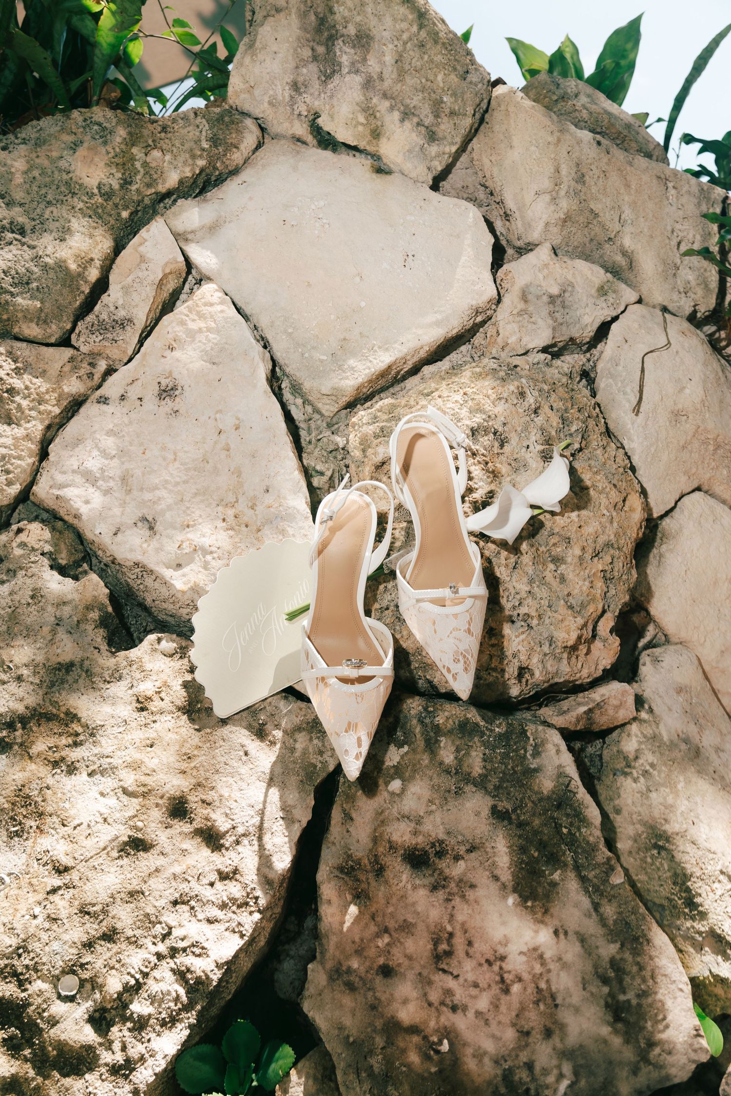 Elegant white lace bridal heels with ankle straps displayed on natural stone rocks surrounded by green foliage outdoors.